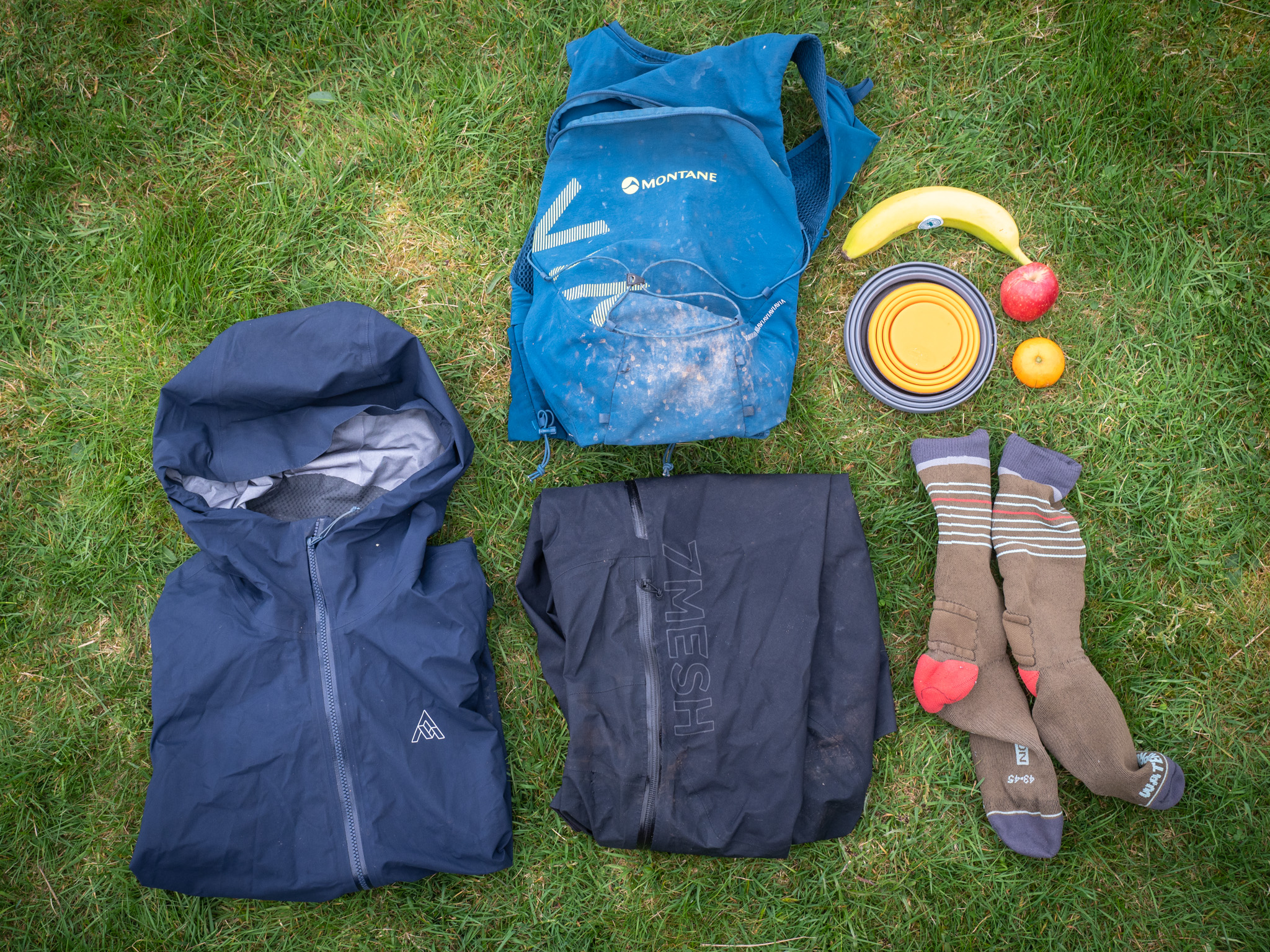 Flat lay on grass of two jackets, a running vest, a folding bowl, fruit and wool socks