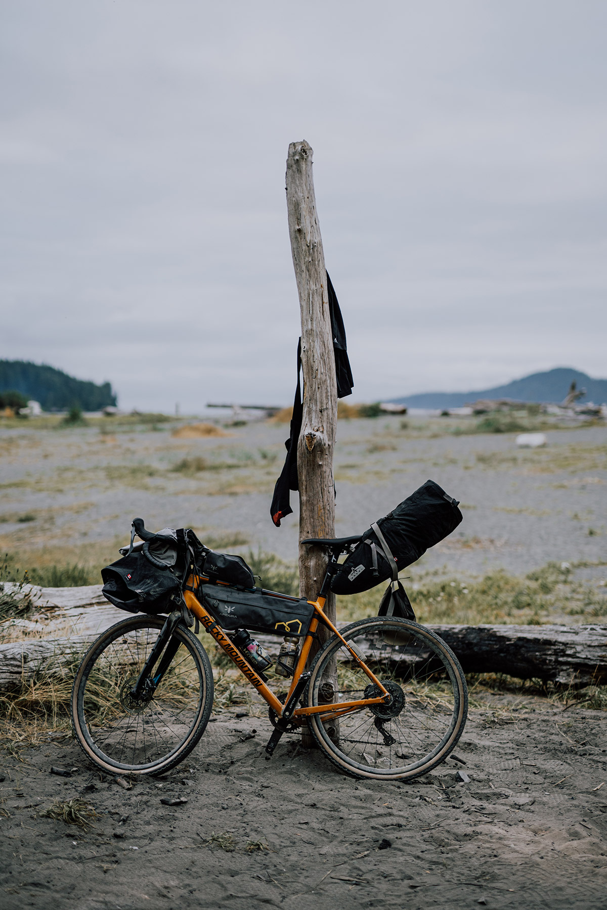 Loaded orange gravel bike leaning on a driftwood post on a grey beach, shorts drying above
