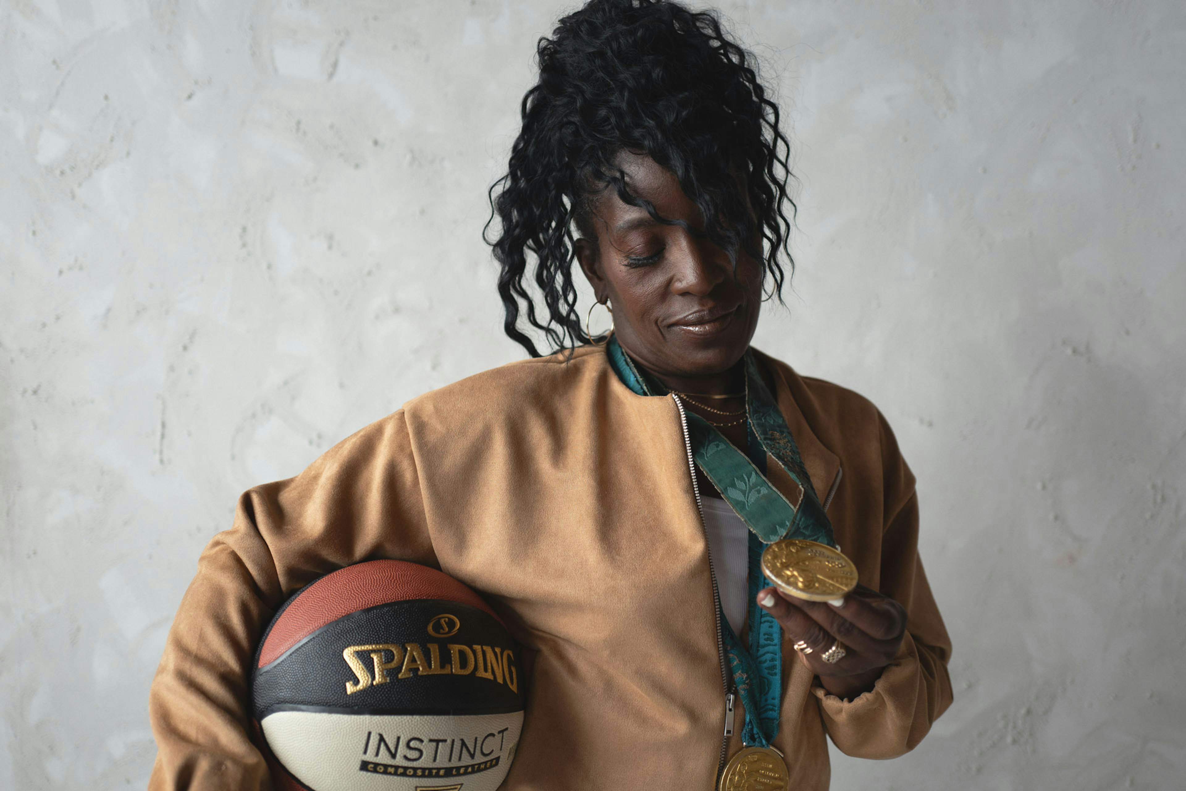 Ruthie Bolton holds a basketball and examines a gold medal while wearing a tan jacket against a textured light-gray wall.