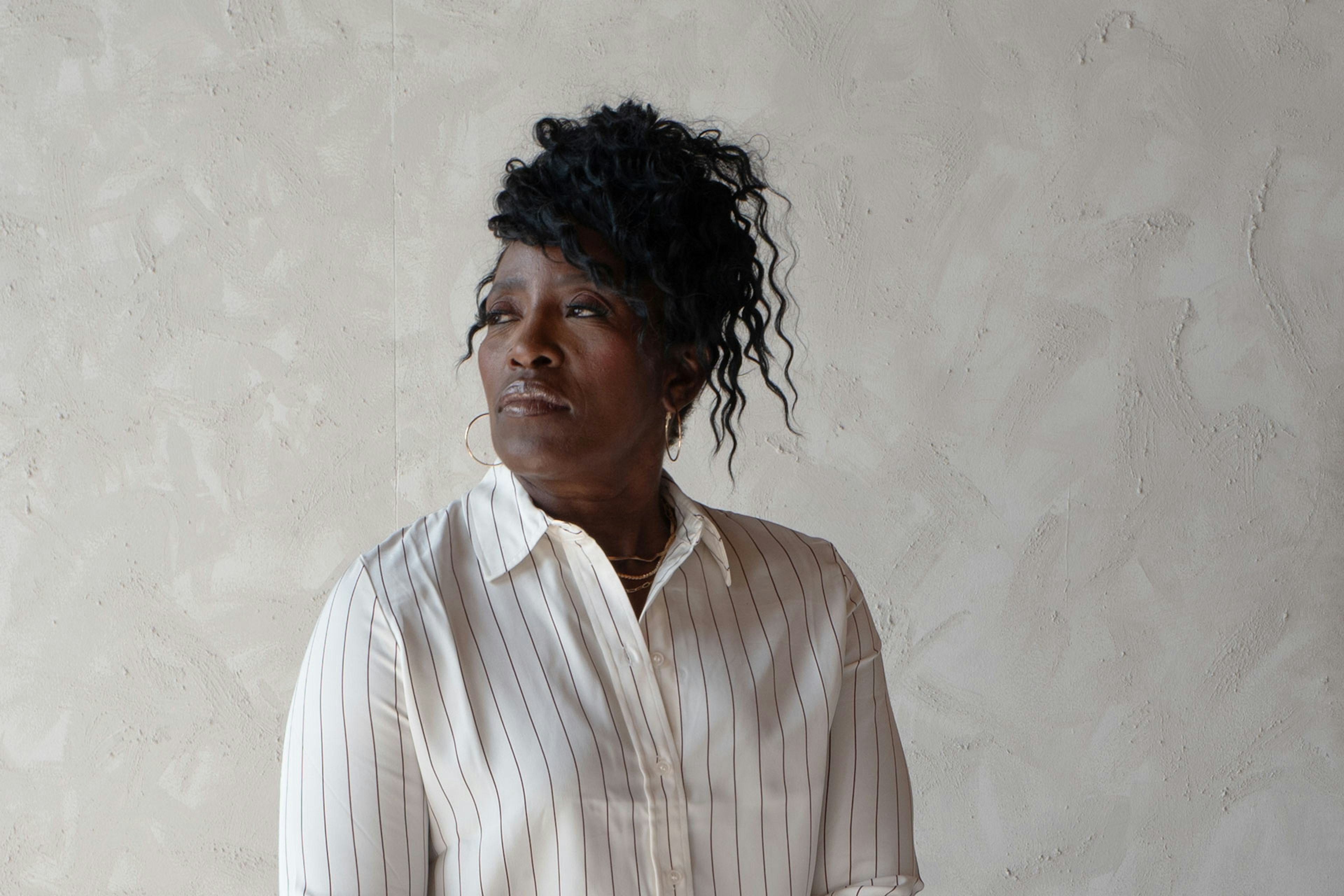 Ruthie Bolton in a white pinstriped blouse looks thoughtfully to the side against a textured light-gray wall.