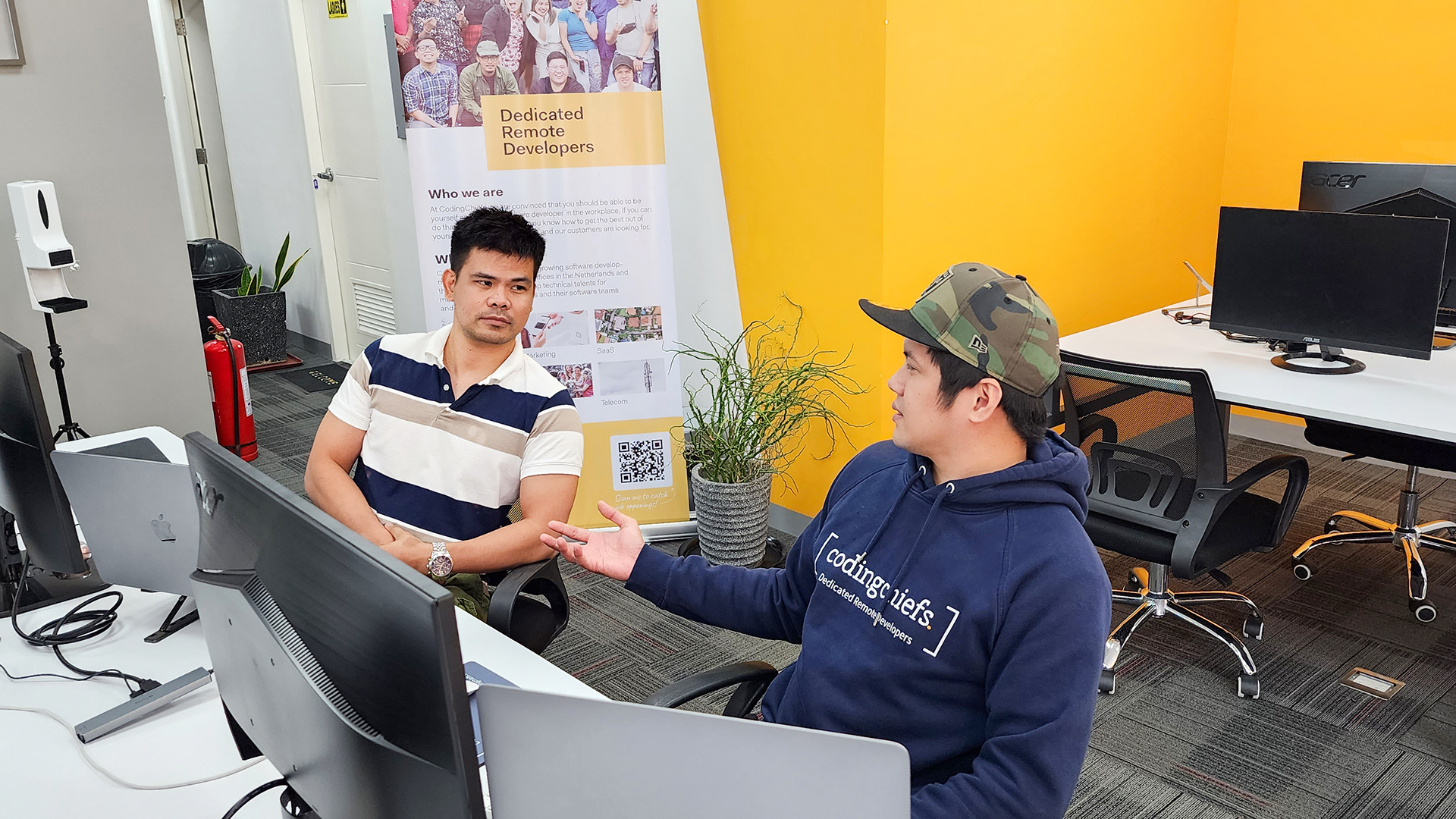 Two CodingChiefs developers talking at their desks, one in a CodingChiefs hoodie, beside a Dedicated Remote Developers banner