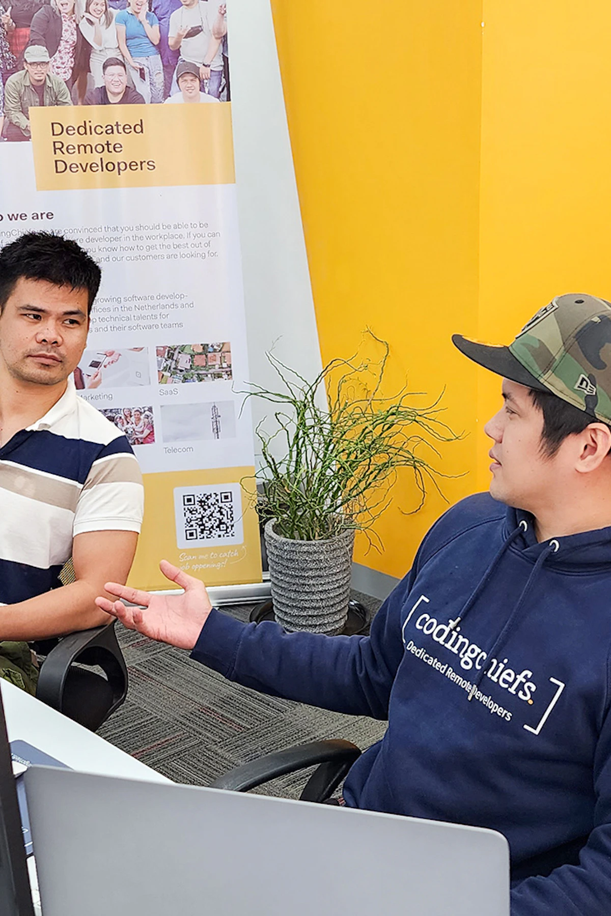 Two CodingChiefs developers talking at their desks, one in a CodingChiefs hoodie, beside a Dedicated Remote Developers banner