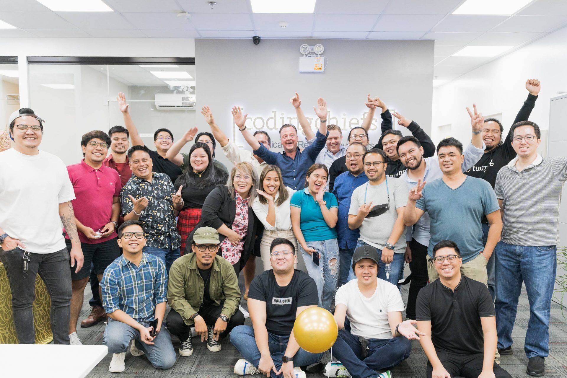The CodingChiefs team cheering with their arms raised in front of the office wall logo