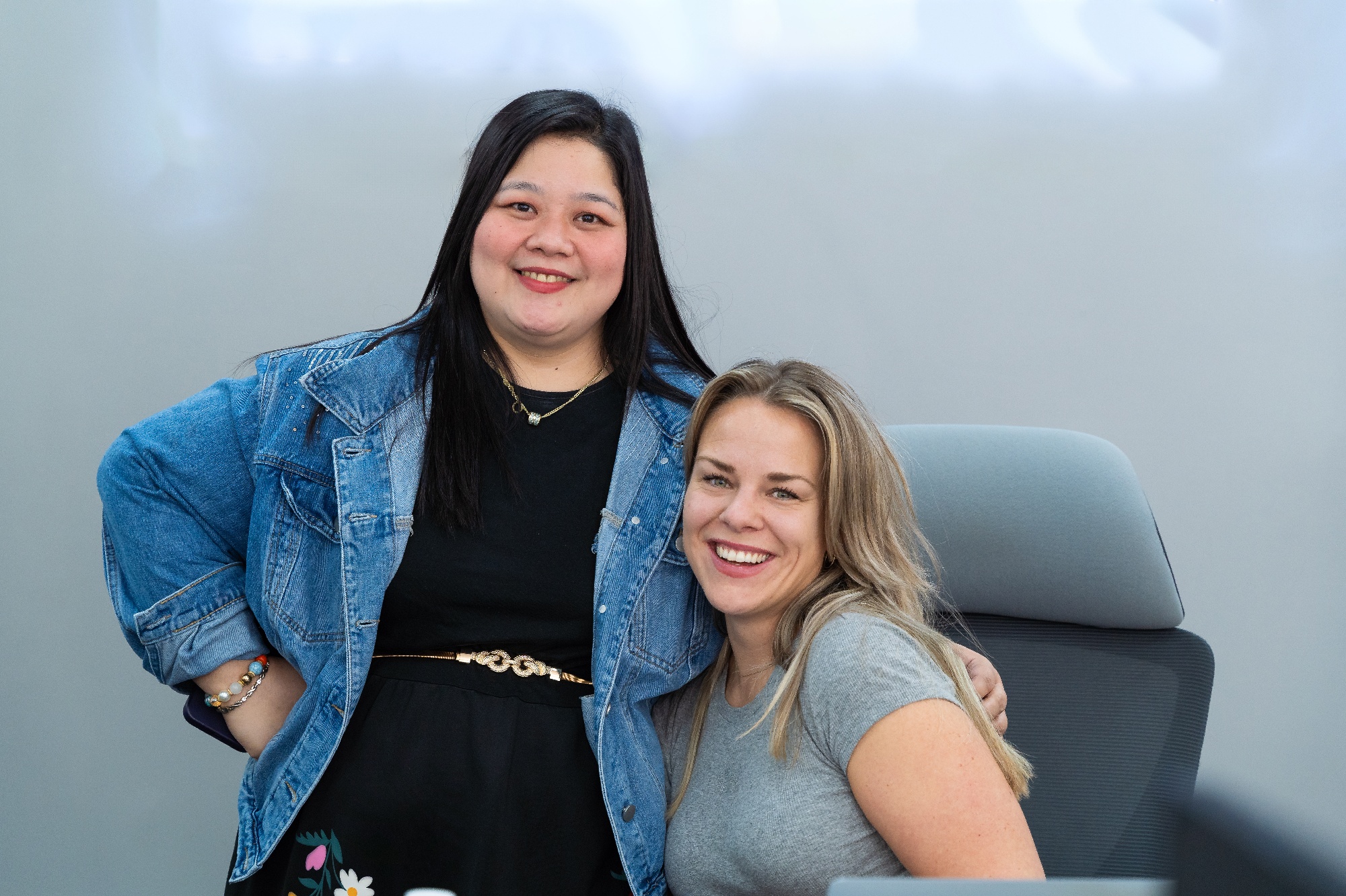 Two CC.Talent colleagues smiling at the camera in the office, one standing in a denim jacket with her hand on her hip and the other seated at her desk in a grey T-shirt