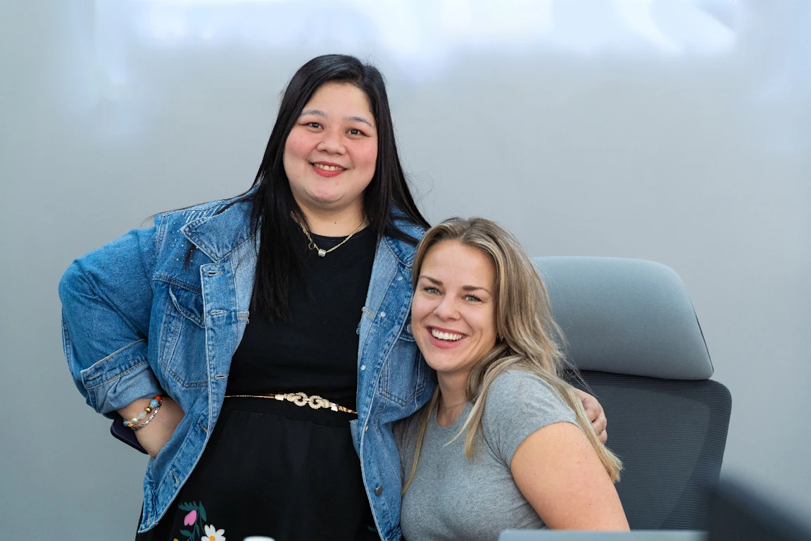 Two CC.Talent colleagues smiling at the camera in the office, one standing in a denim jacket with her hand on her hip and the other seated at her desk in a grey T-shirt