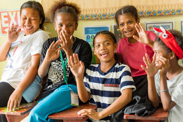 Five schoolgirls laughing and waving together at a classroom desk