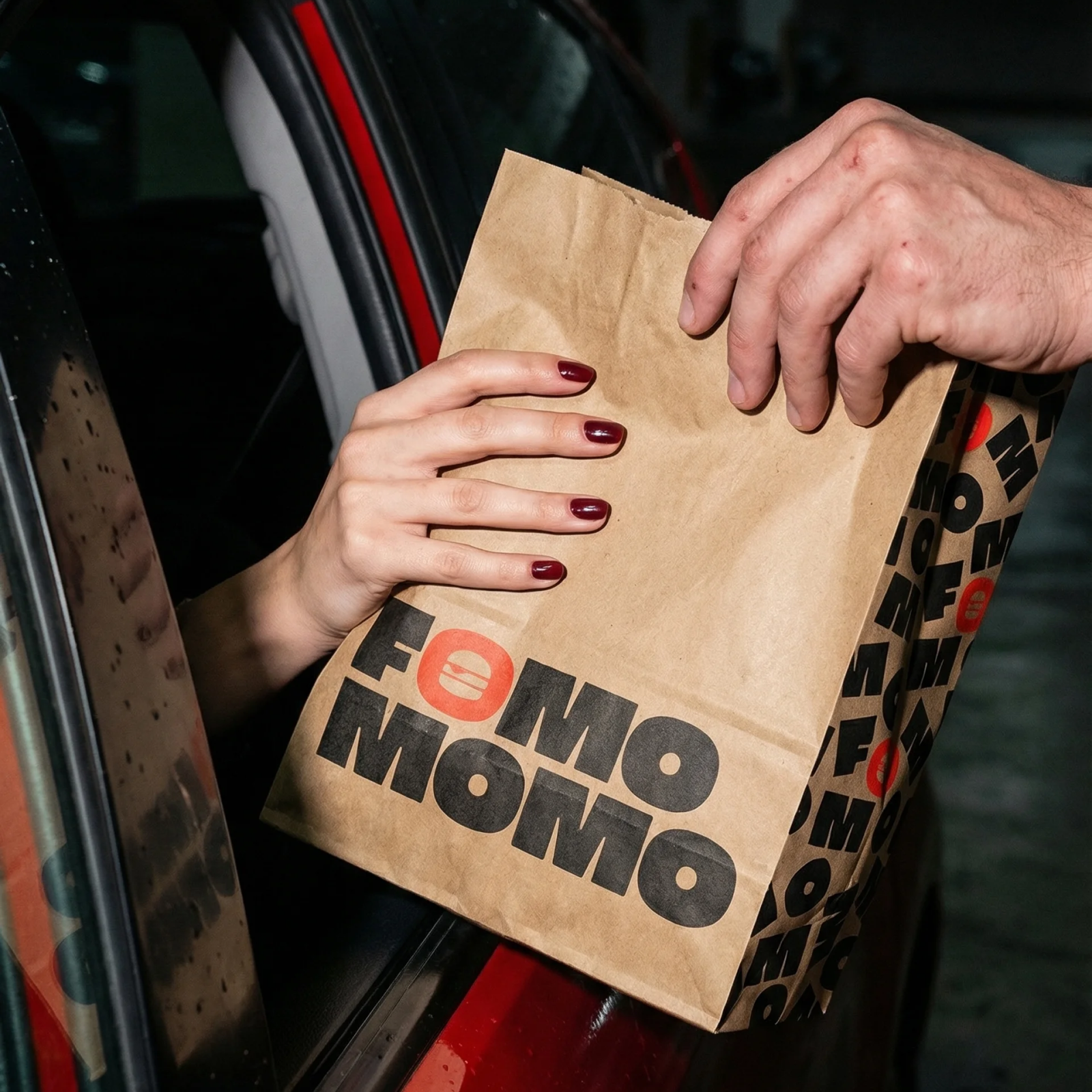 A woman's hand reaching outside a car window grabbing a brown paper take out bag being handed to her. Fomo Momo branding all over the front and sides of the bag