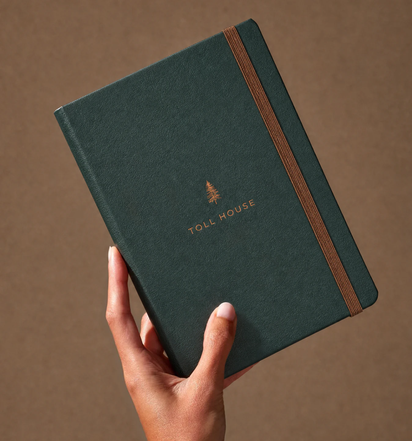 A person holding a toll House branded notebook with pine tree icon and elastic closure in front of a darker beige brown background