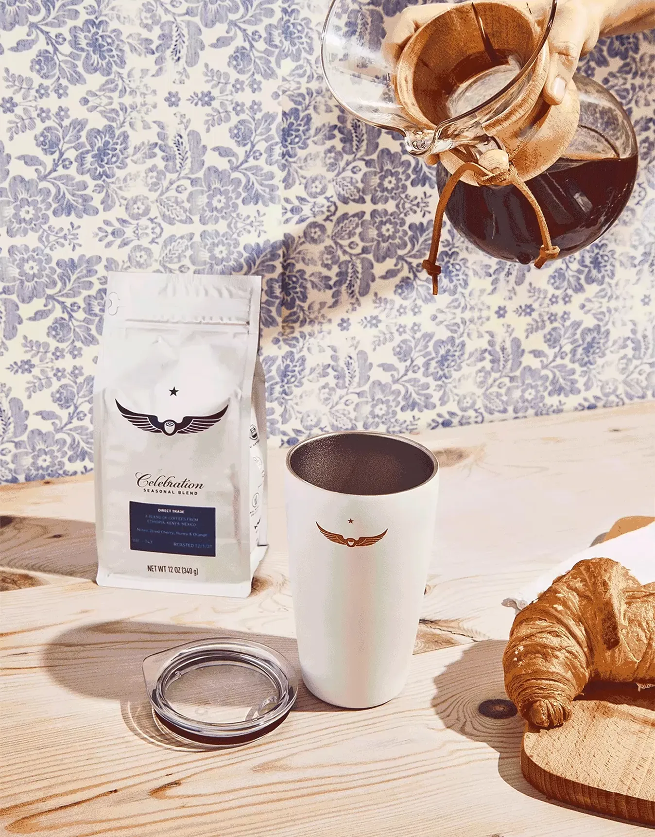 Coffee being poured into a white tumbler beside a Celebration Blend bag and croissant on a wood table