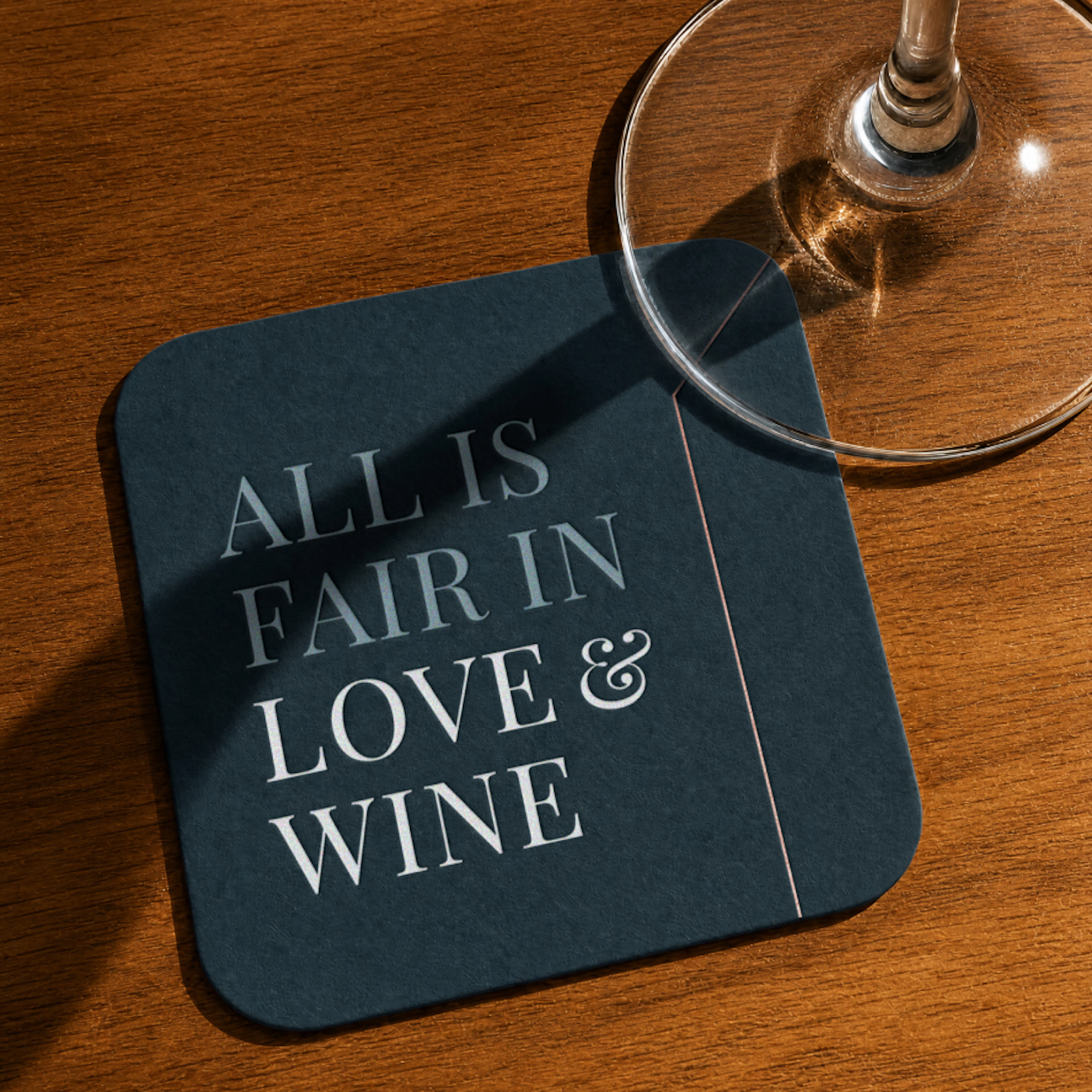 Top down view of a dark blue square coaster that says "All is fair in love & wine" on the top right corner is a wine glass sitting on the coaster