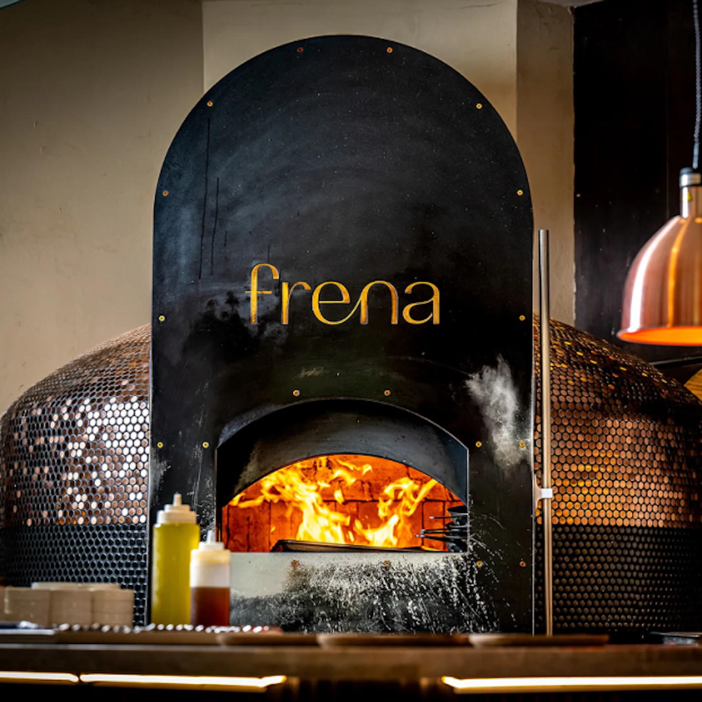 Frena wordmark on a black wood-fired oven with flames inside