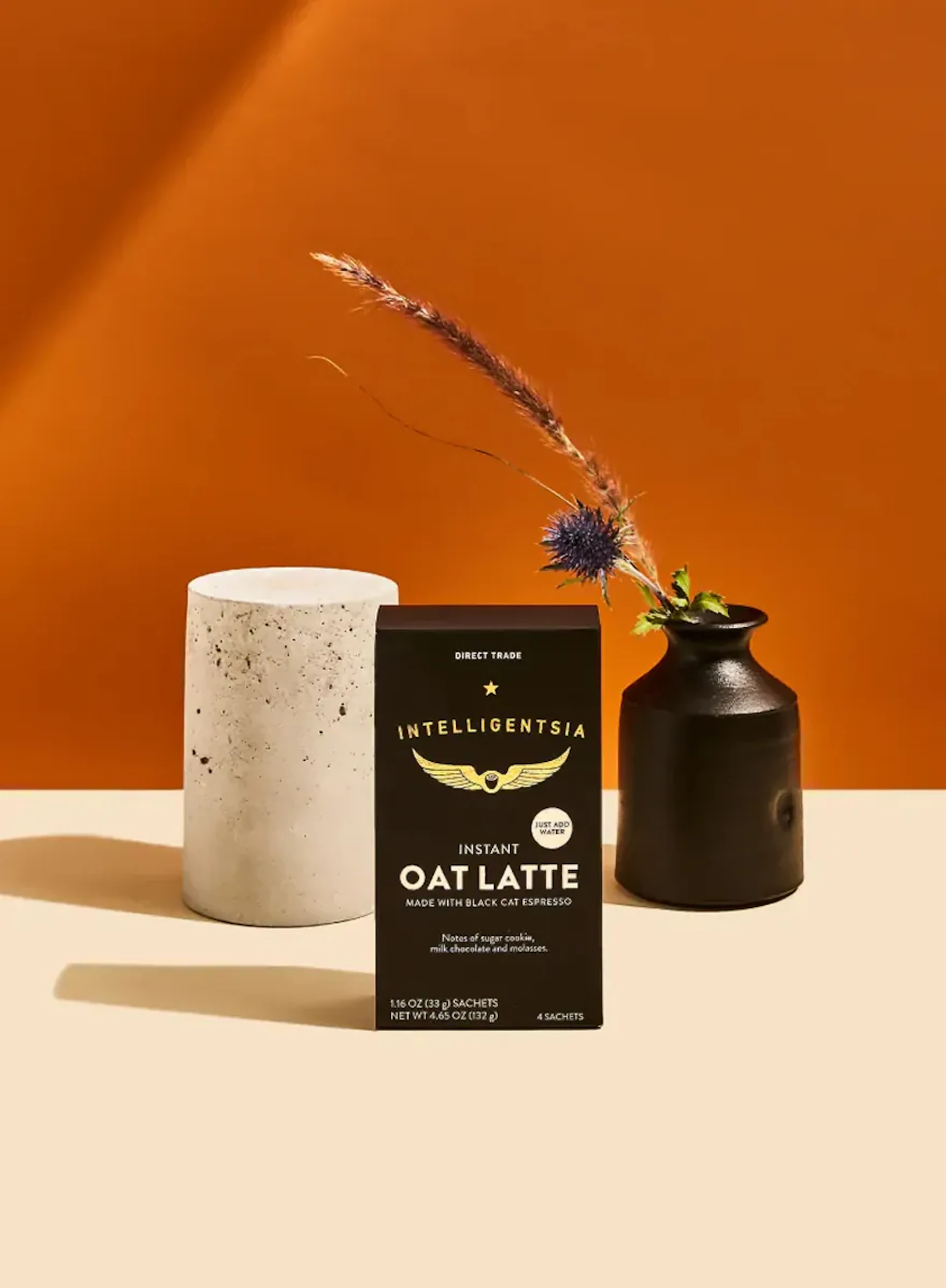 Intelligentsia Instant Oat Latte box beside a concrete cylinder and dark ceramic vase on an orange background