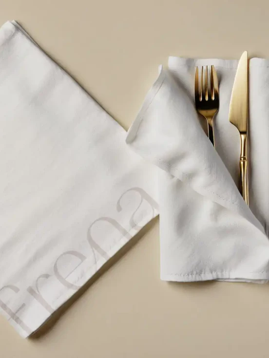 Top down view of a fork and knife sitting with a white napkin with another white napkin on the left with the Frena logo printed in grey