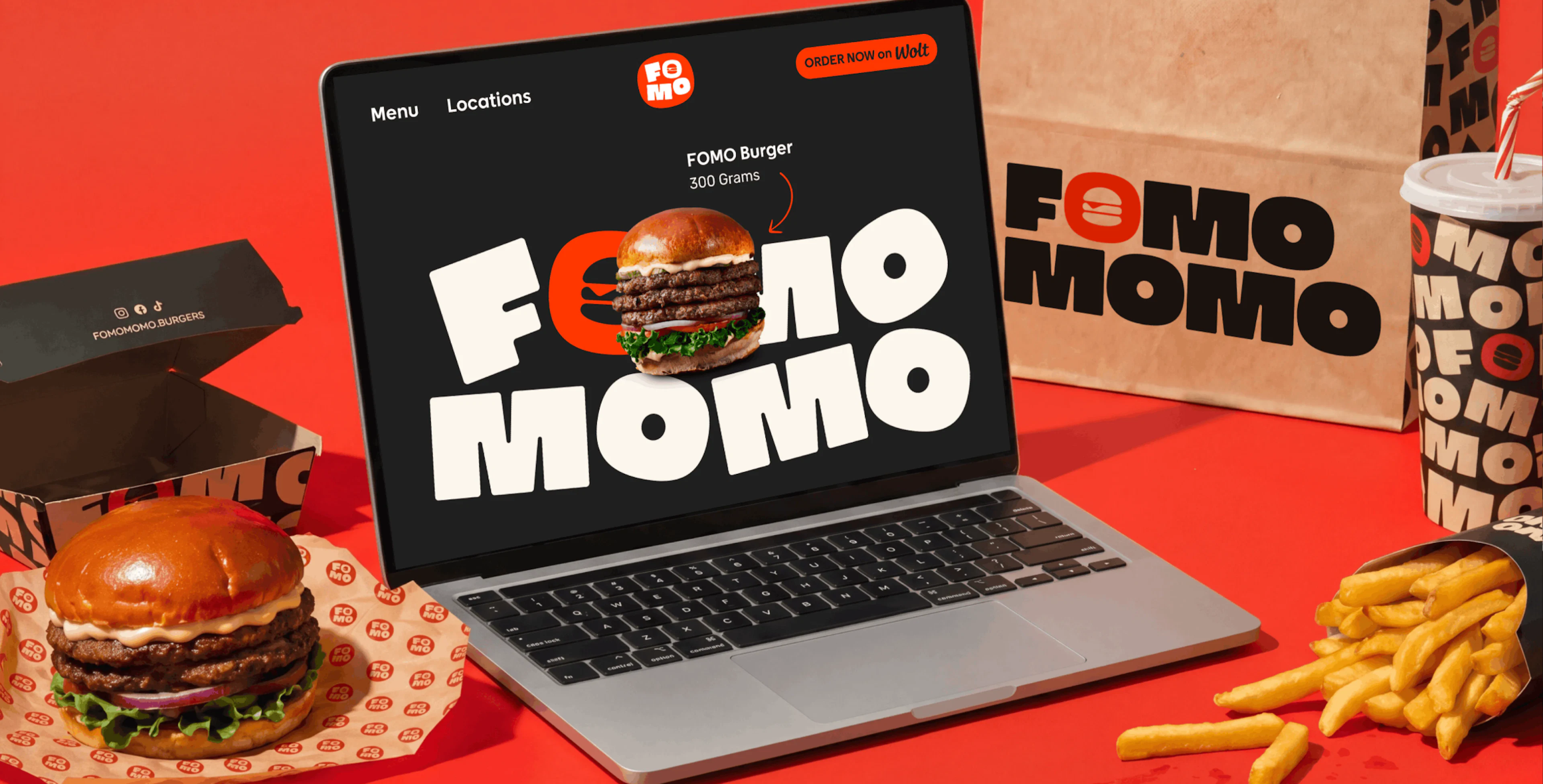 A laptop displaying the Fomo Momo website and a burger image, surrounded by branded paper bags and food items on a red background.