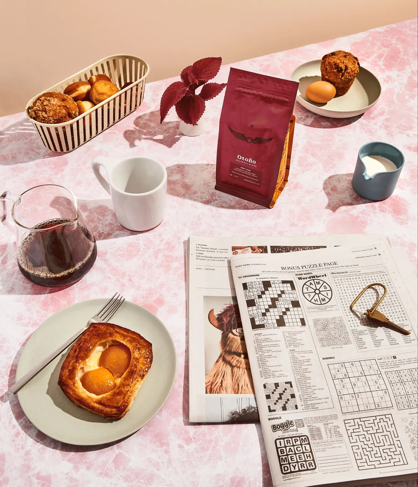 Overhead shot of a burgundy Otoño coffee bag on a pink marble table with pastries, coffee, and a newspaper puzzle page