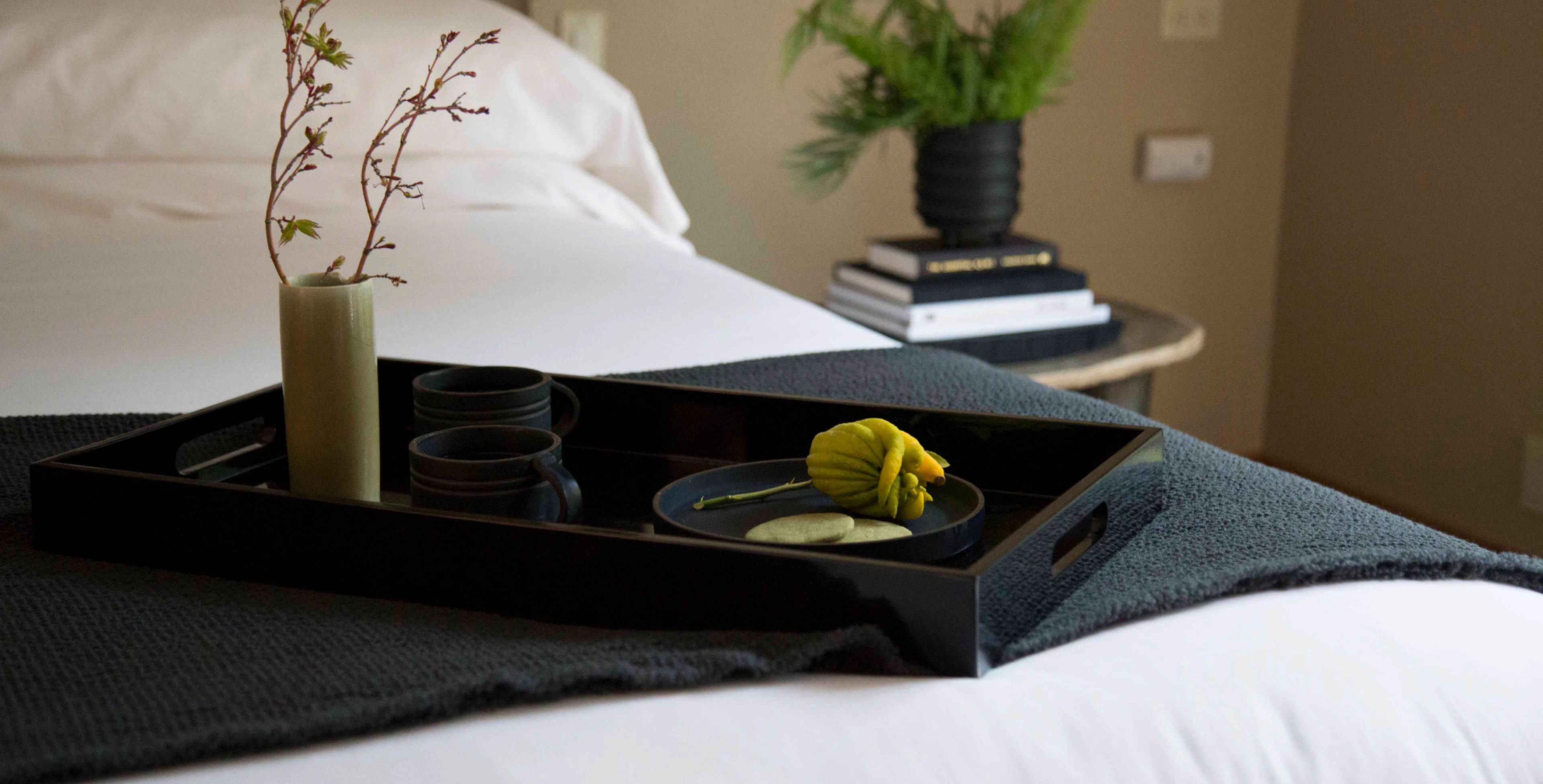 A black wooden tray sitting on top of a modern hotel bed with white comforter and pillows. The tray has a minimal plant, and printed materials