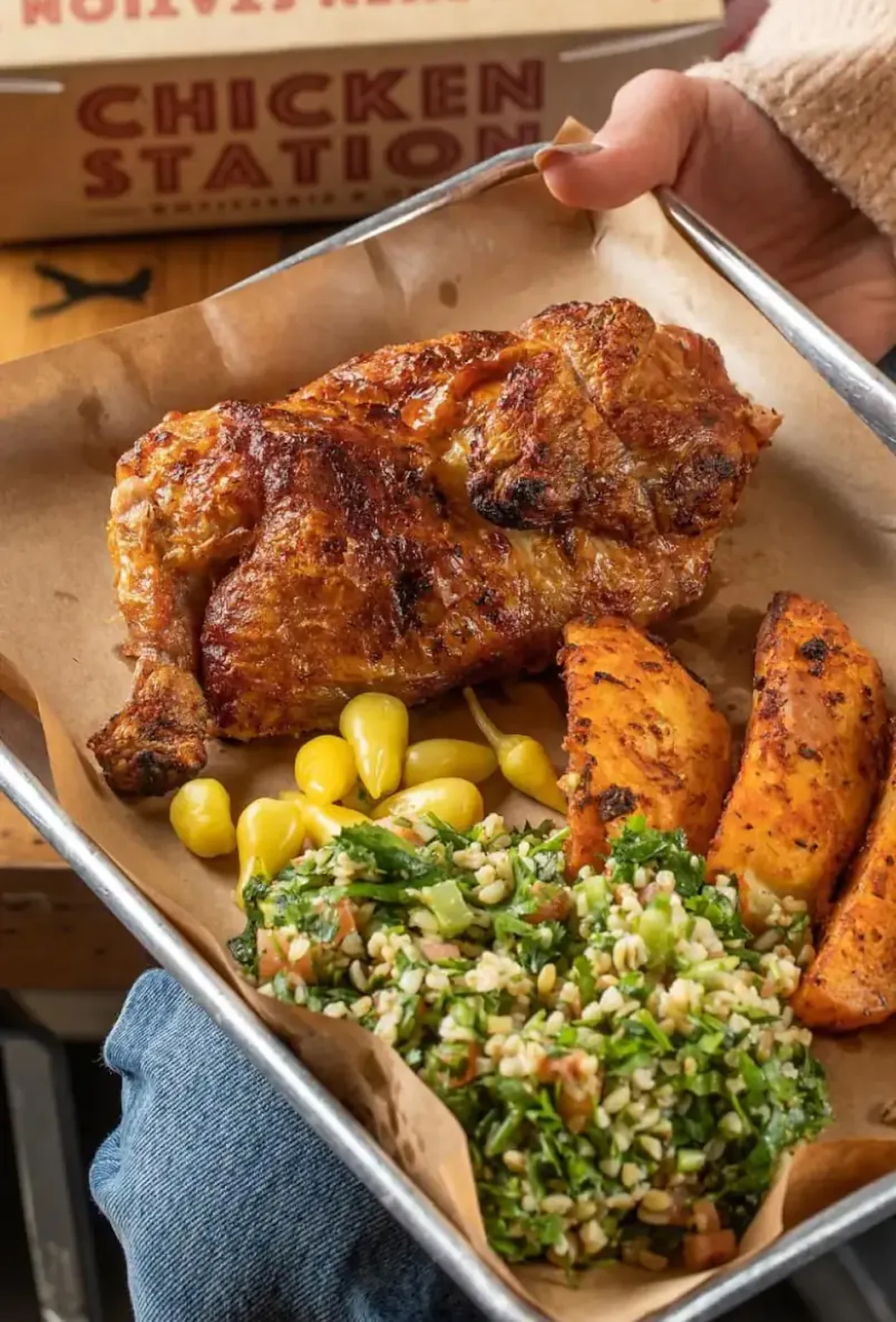 Hand holding a tray of grilled chicken, corn, and tabbouleh salad