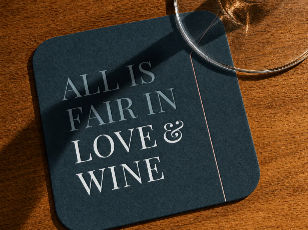 Top down view of a dark blue square coaster that says "All is fair in love & wine" on the top right corner is a wine glass sitting on the coaster