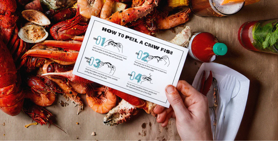 A top down view of different seafoods like crap, lobster and shrimp arranged on a table, with a person's hand holding a card that describes how to peel a craw fish with instructions and illustrations