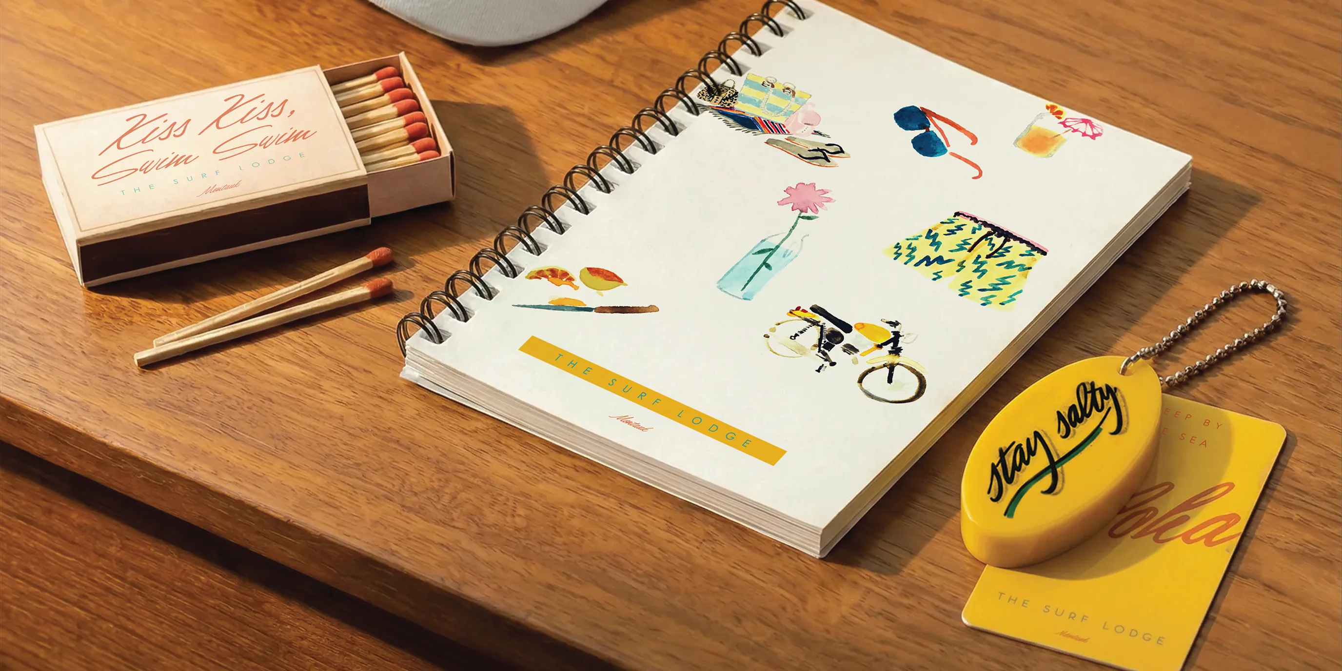Matchbox, spiral notebook with illustrations, and yellow keychain on a wood table