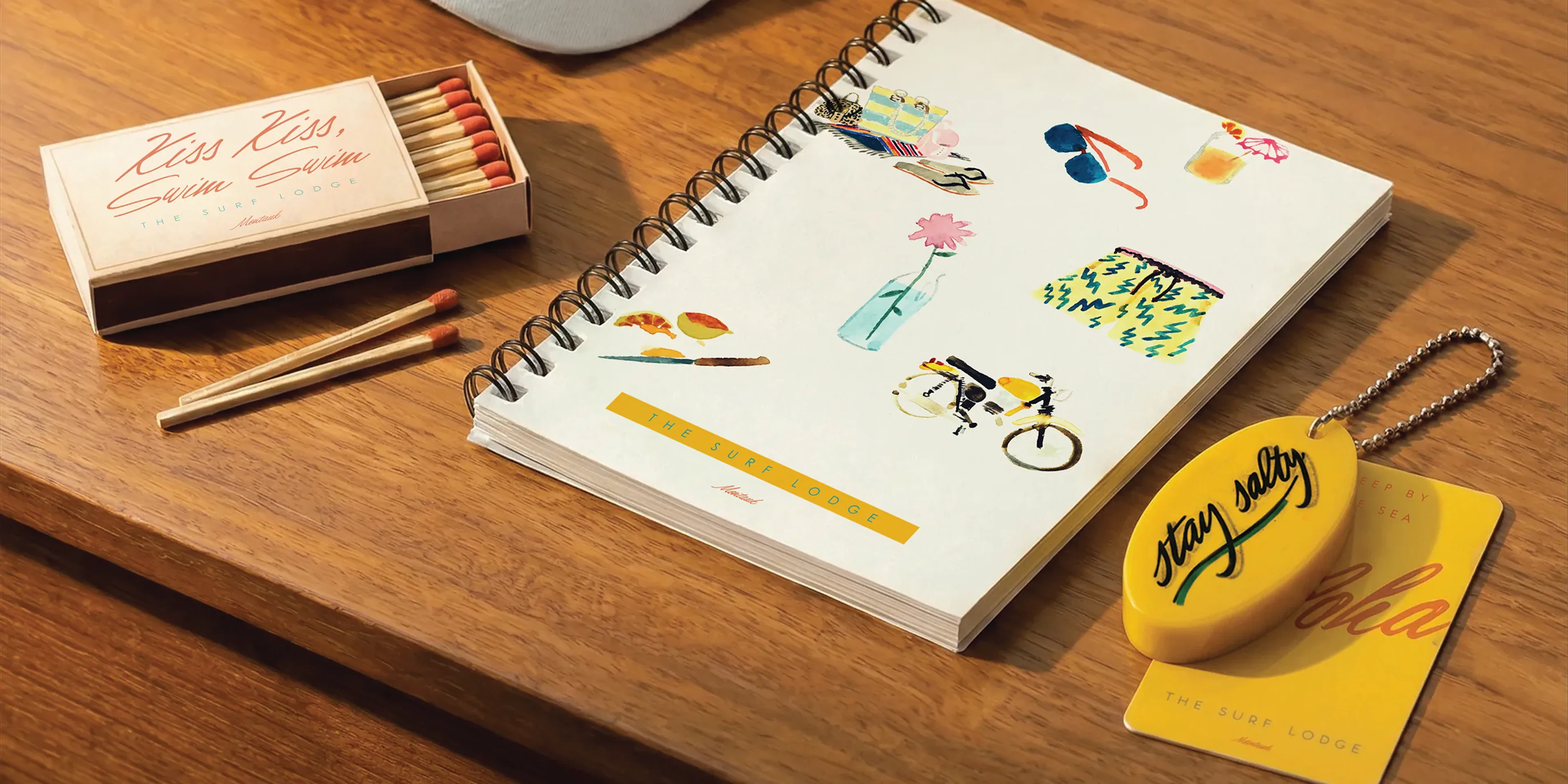 Matchbox, spiral notebook with illustrations, and yellow keychain on a wood table