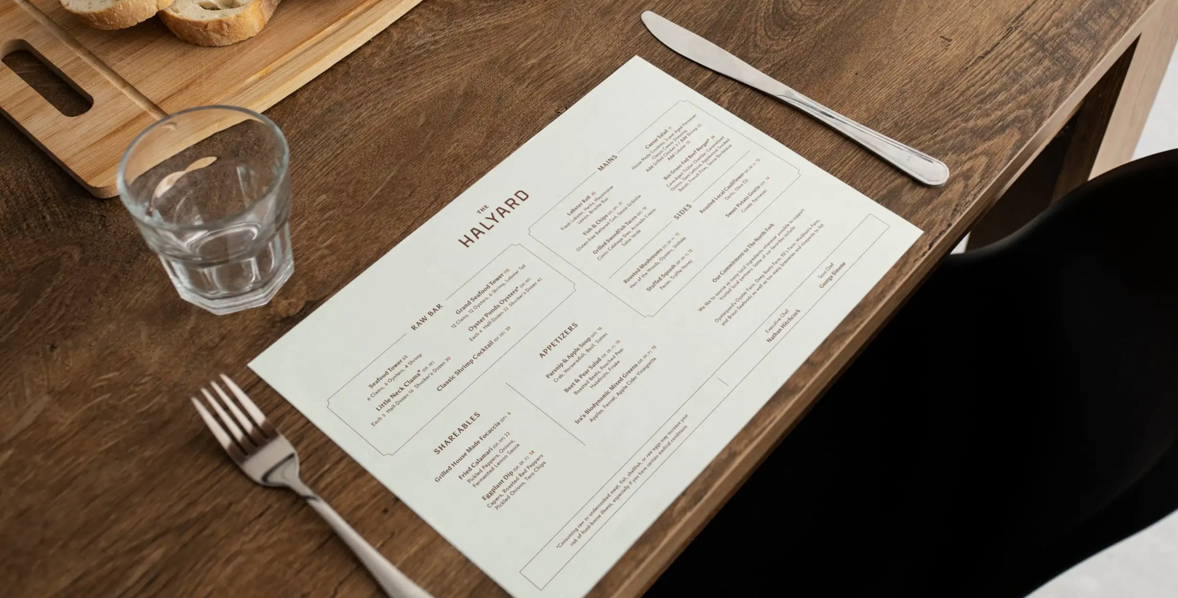 A top down view of a menu sitting on a dark wood table with cutlery on either side, bread is on the table as well as a water glass
