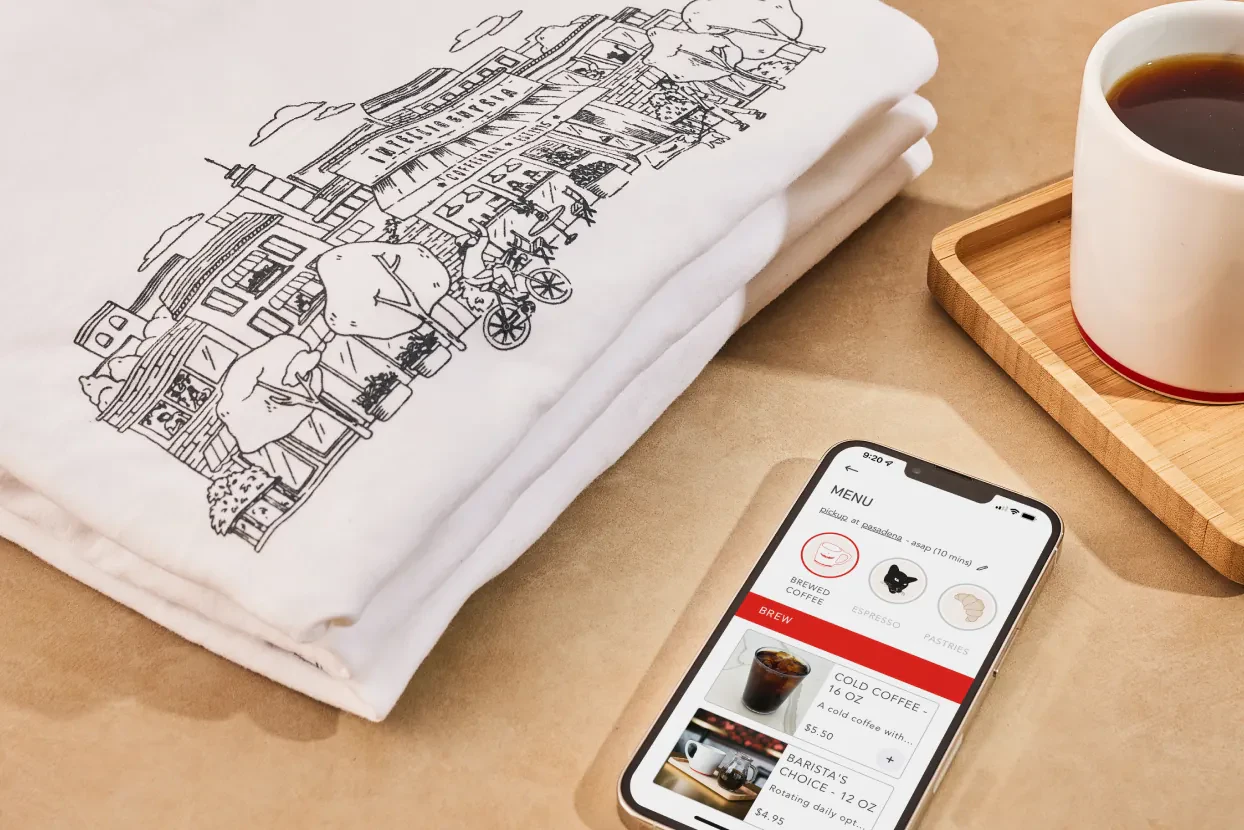 Folded illustrated t-shirt beside a phone showing a menu app and coffee cup
