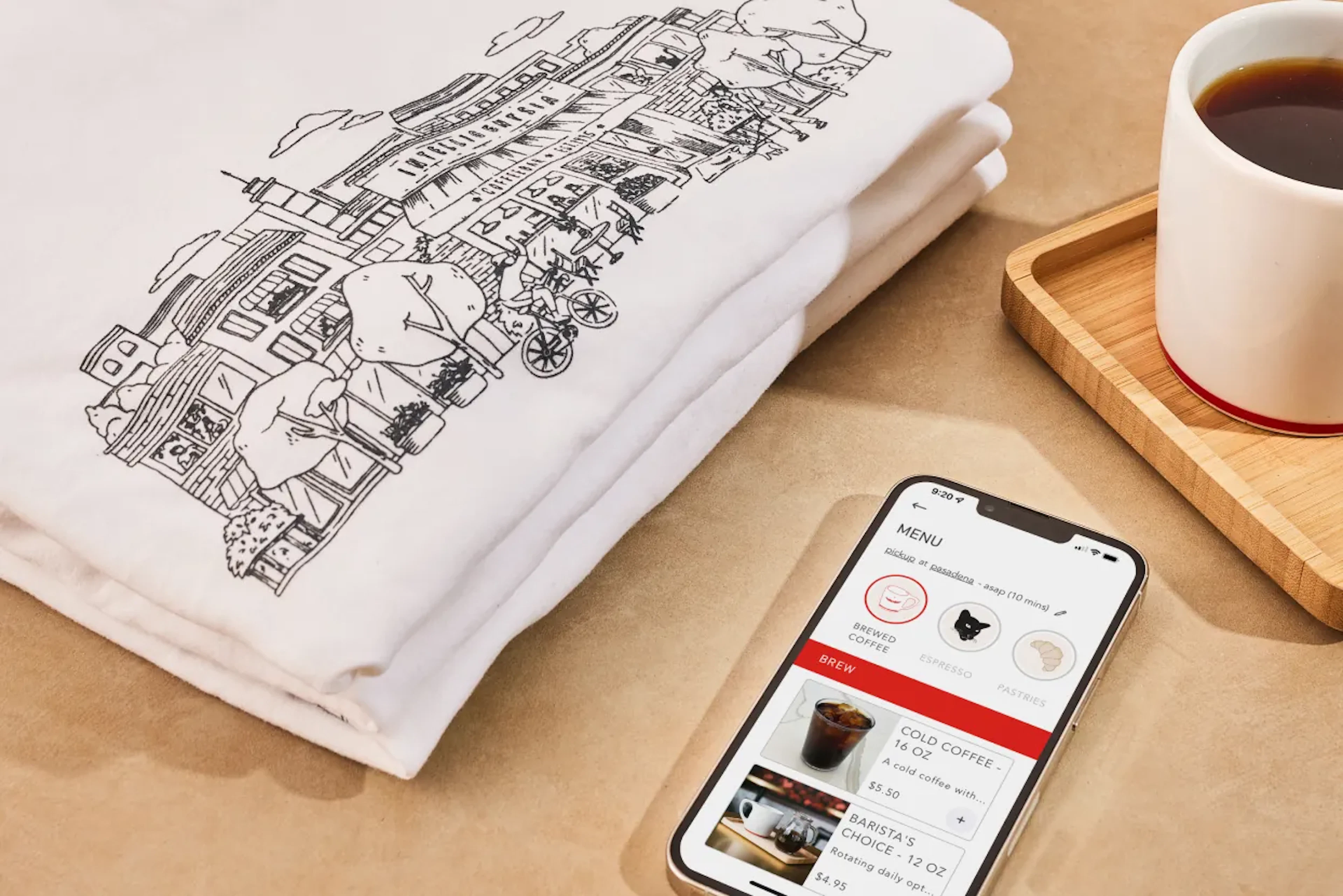 Folded illustrated t-shirt beside a phone showing a menu app and coffee cup