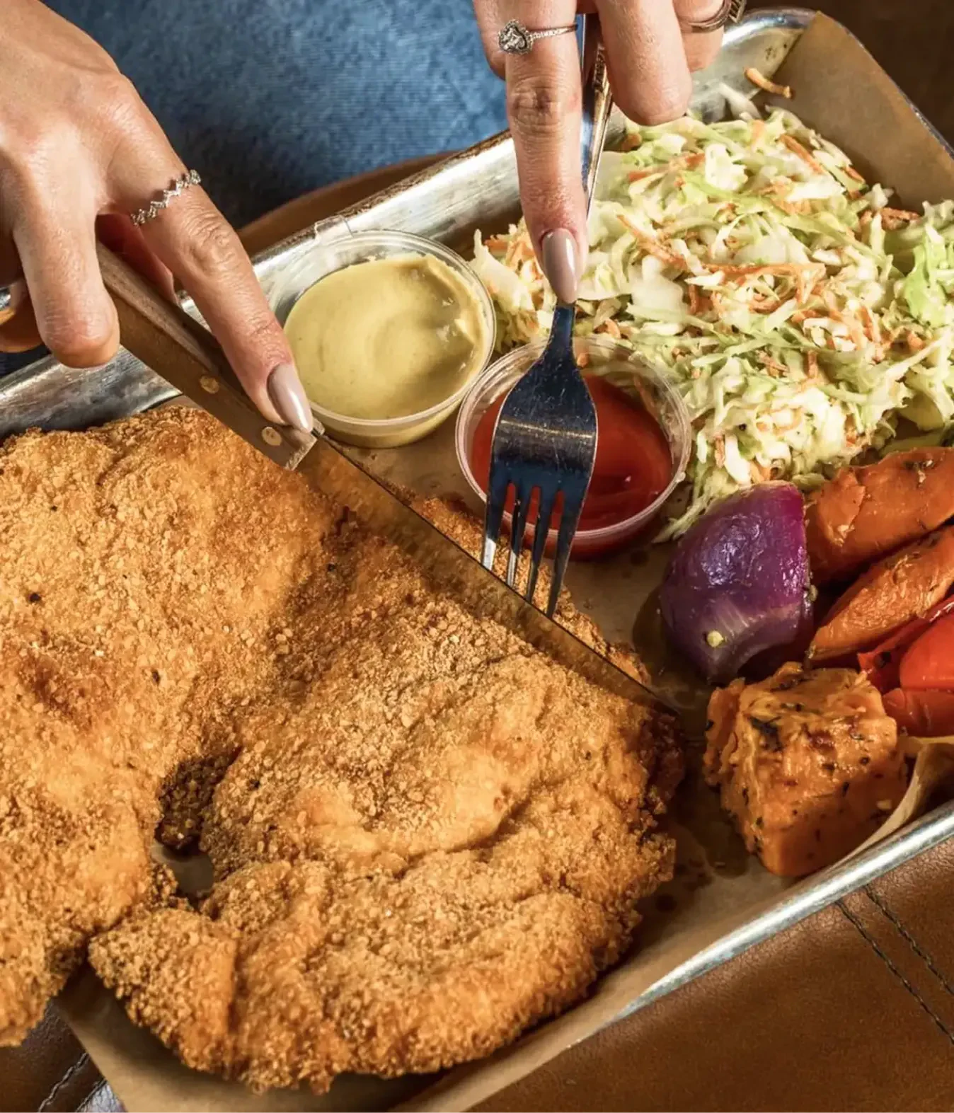 Hands cutting a breaded chicken schnitzel with coleslaw and sides