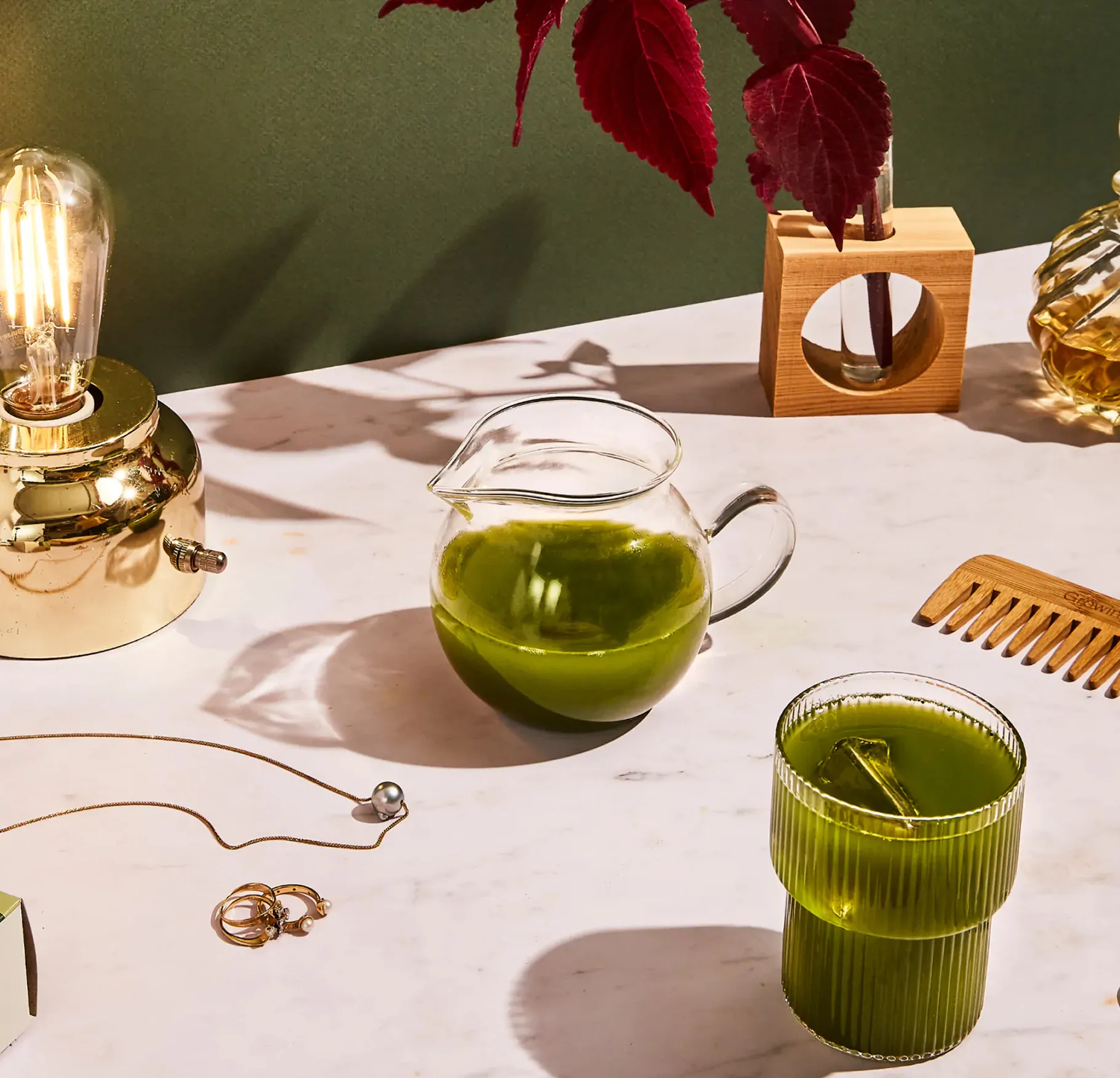 Green tea in a glass pitcher and ribbed cup on a marble table with a brass lamp and jewelry