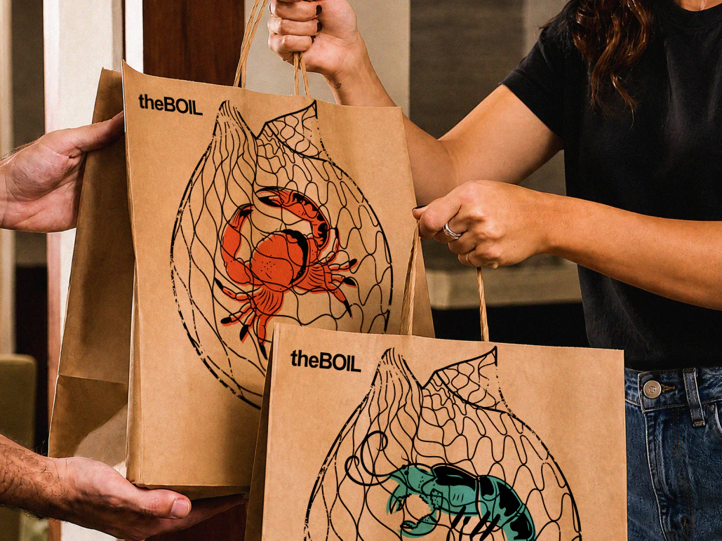 Woman taking 2 brown paper takeout bags from a delivery person, the front of the bags have the boil logo and crab and shrimp tattoo style illustrations