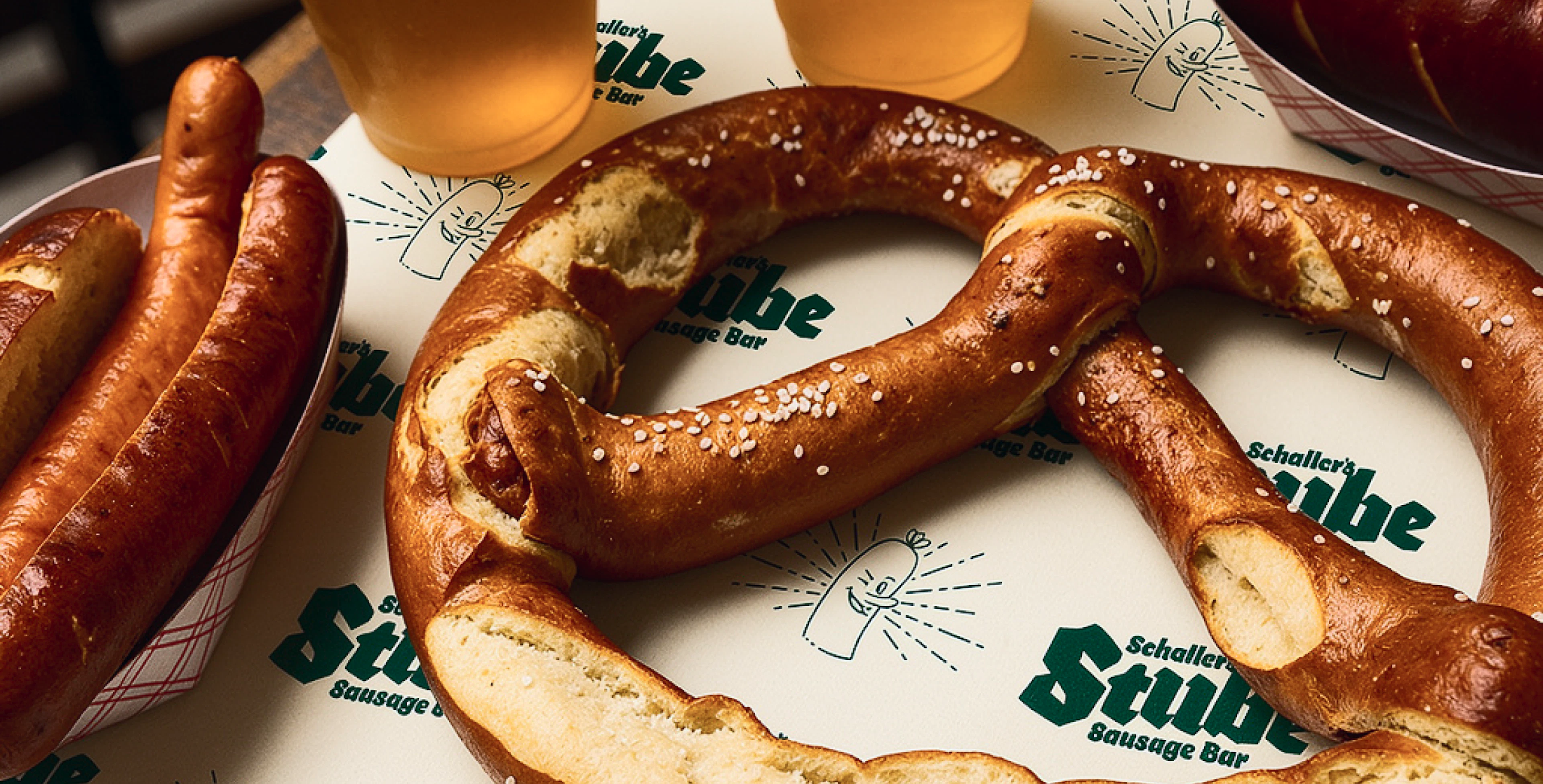 A large round golden-brown sesame-topped bagel surrounded by hotdogs and beer and repeated green Stube logo on light beige paper tray