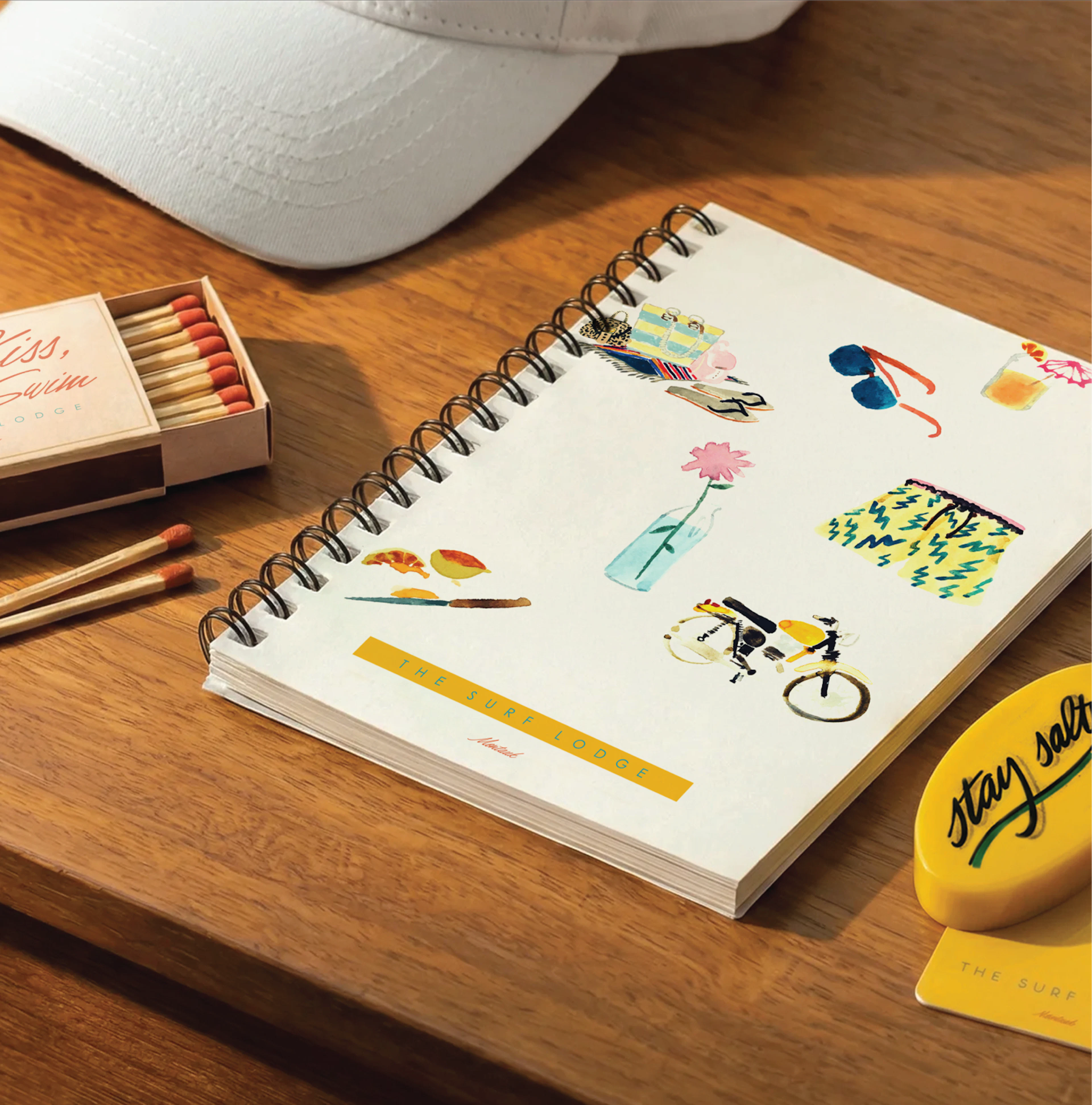 A flat lay of creative stationery on a warm wooden surface, featuring a white spiral-bound notebook decorated with colorful hand-drawn illustrations, abstract shapes, and black ink mushroom doodles. A white textured pen, two small boxes of colored pencils, and a yellow branded sticker complete the artistic arrangement.