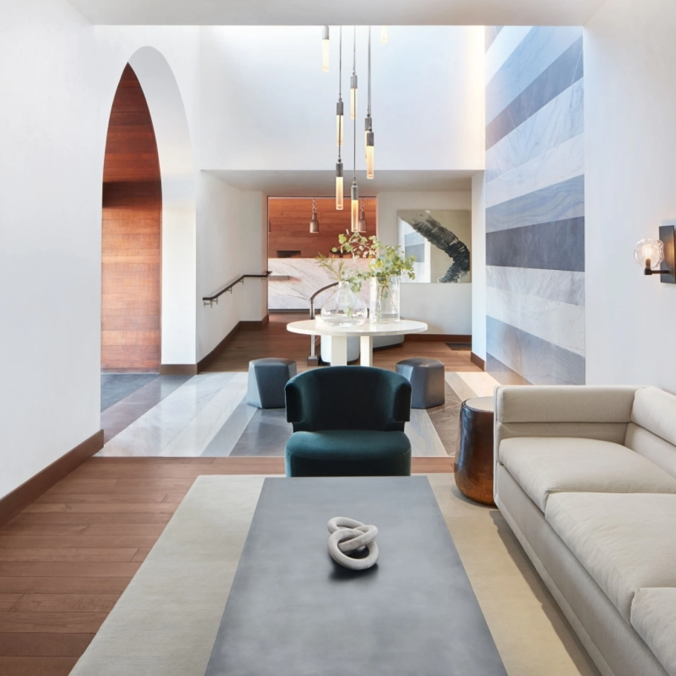 A stylish interior hotel room showcasing the living room with a modern white couch and rectangular coffee table, you can also see into the dining room which has teal velvet chairs and an elegant chandelier