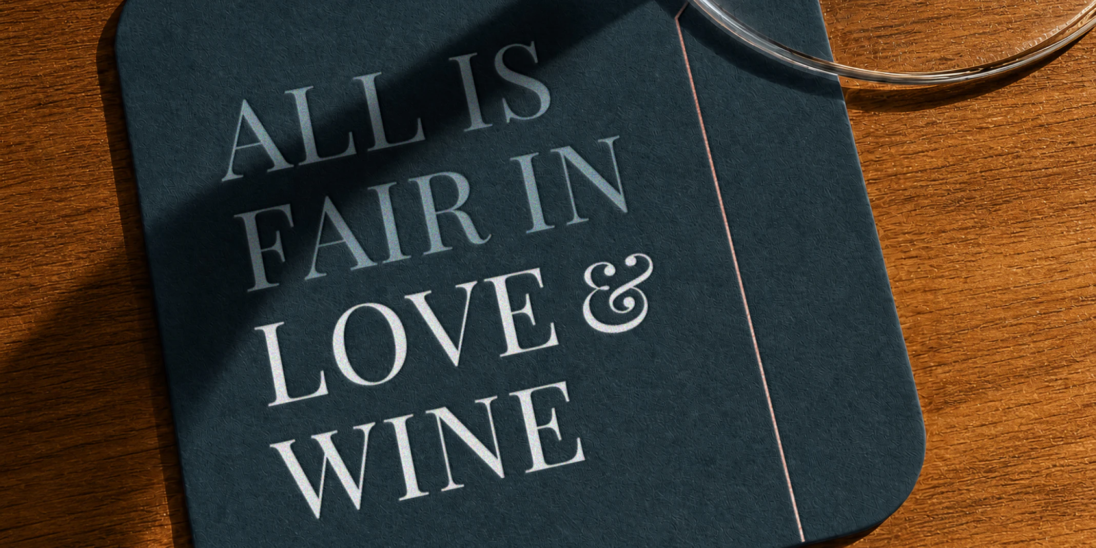 Top down view of a dark blue square coaster that says "All is fair in love & wine" on the top right corner is a wine glass sitting on the coaster