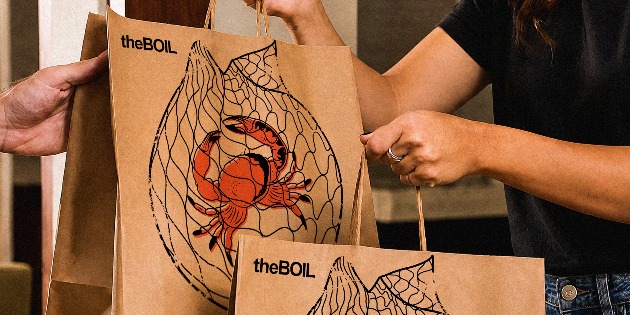 Woman taking 2 brown paper takeout bags from a delivery person, the front of the bags have the boil logo and crab and shrimp tattoo style illustrations