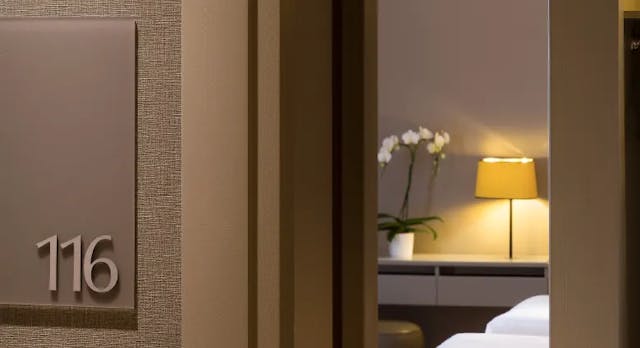Détail, entrée de chambre d'hôtel avec signalétique numéro de chambre et vue de la porte entrouverte sur celle-ci, du lit d'un luminaire et d'une orchidée.