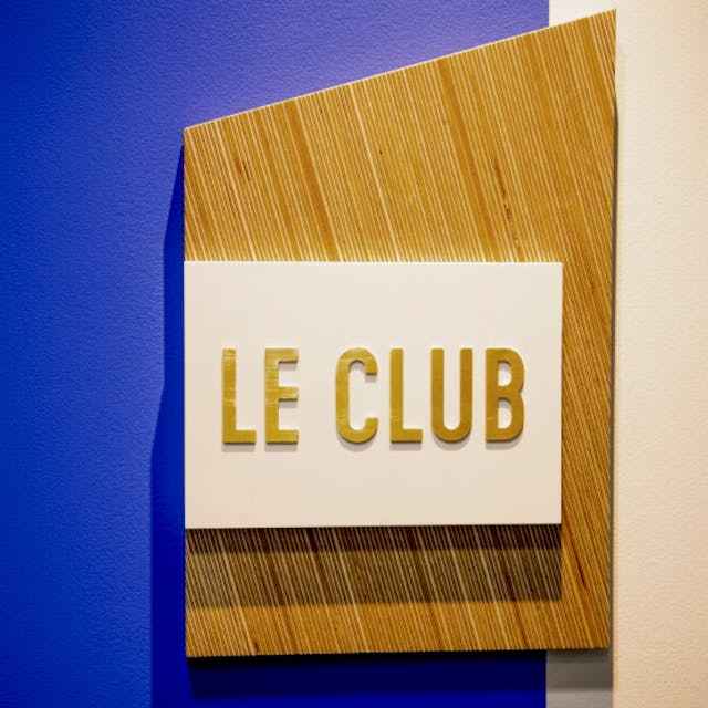 Détail signalétique Le club écrit en lettre doré sur un carré blanc posé sur une planche blanc avec le mur derrière peint en bleu Klein. 