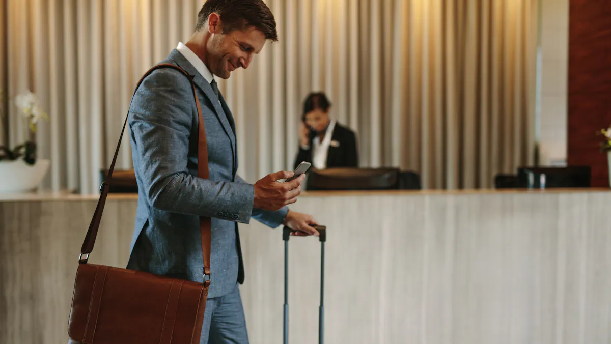 un homme quitte son hôtel avec son ordinateur à l'épaule et sa valise à roulettes en consultant son téléphone portable