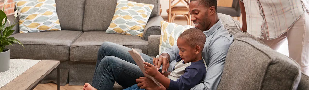 A man sitting on a sofa consults his tablet with a smiling young boy in his arms