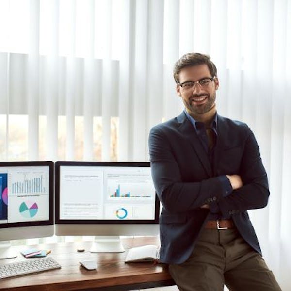 Un homme souriant assis sur une table avec des écrans qui montrent des tableaux de bords de données 
