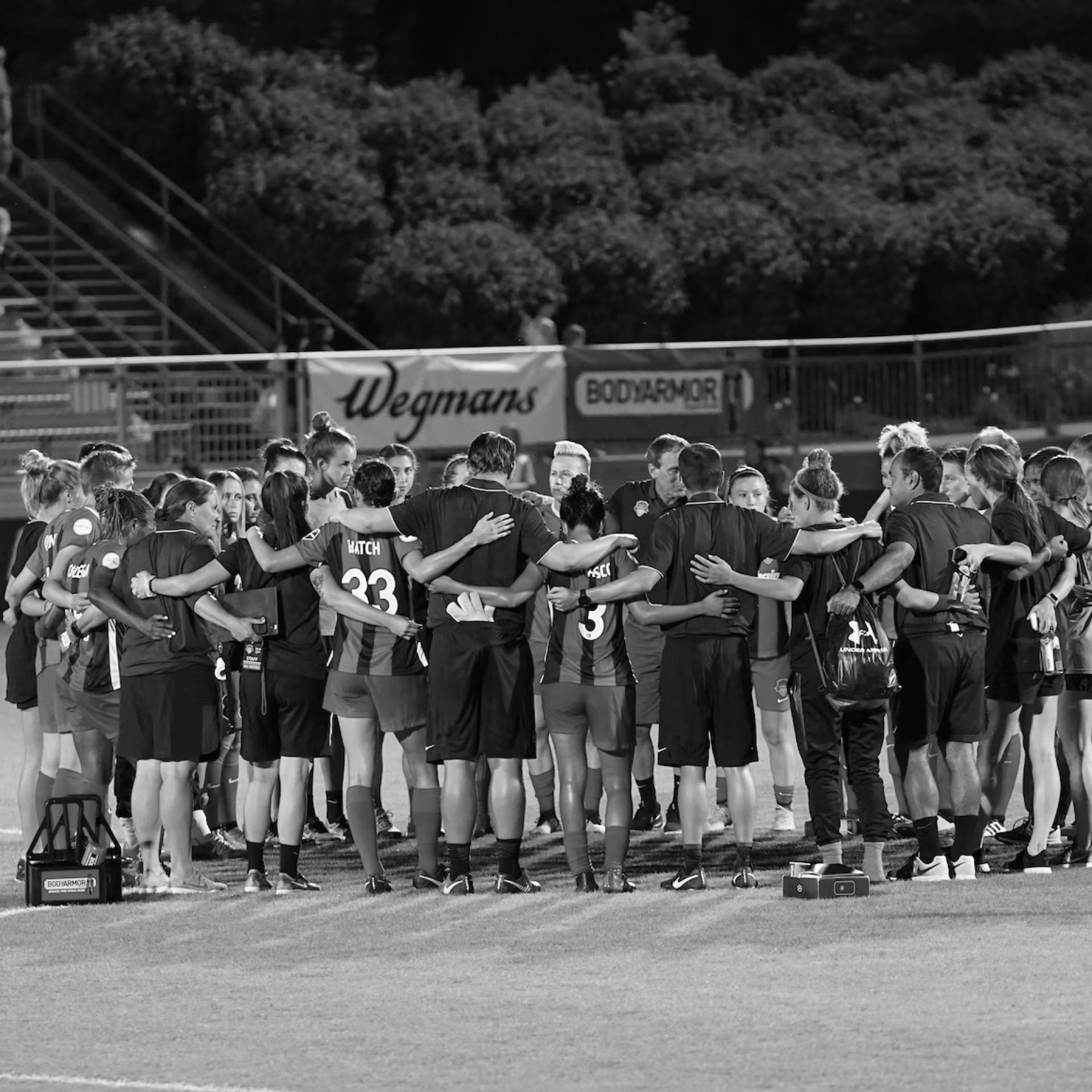 A team of youth football players and their coach huddle up with arms around each other on a soccer field