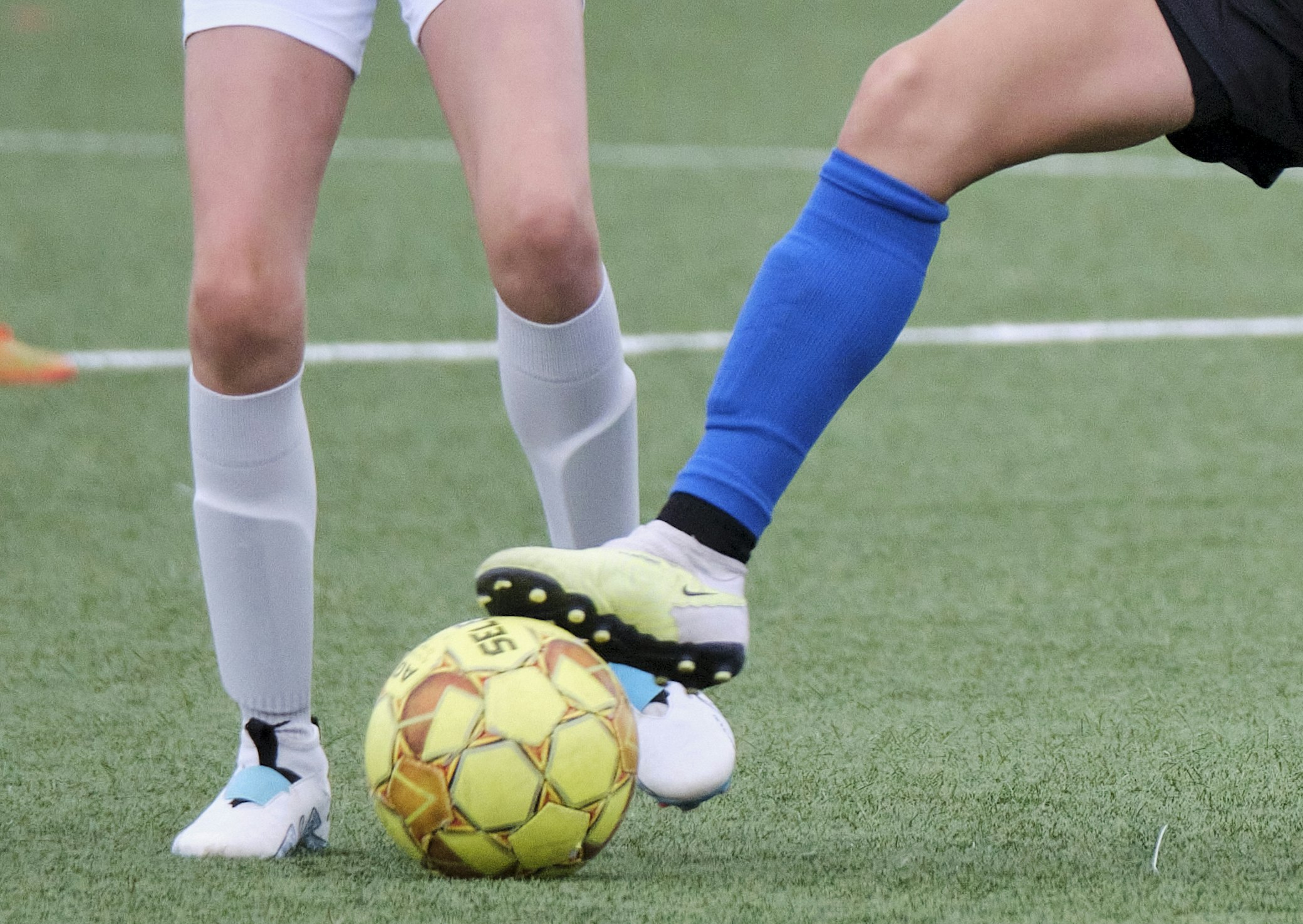 Two players' legs competing for yellow football on artificial turf pitch