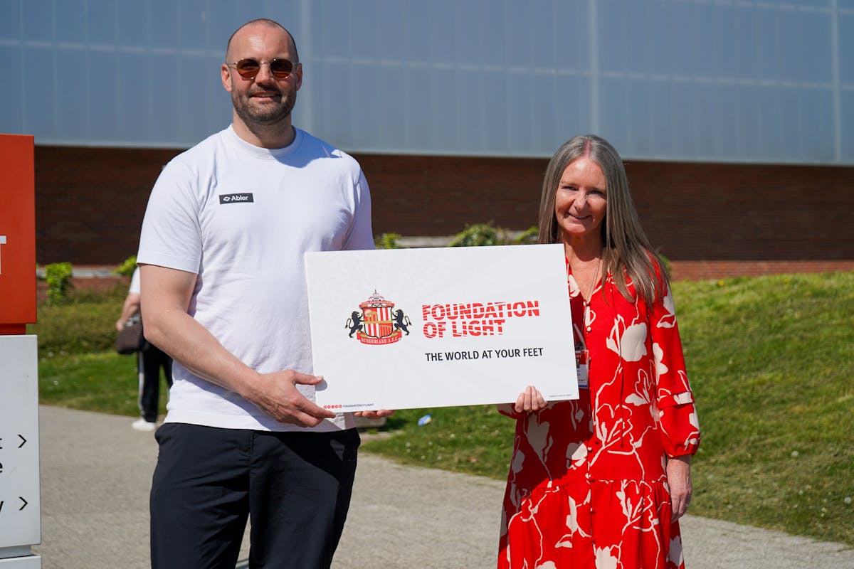 A man in a white t-shirt and a woman in a red floral dress hold a sign that reads "FOUNDATION OF LIGHT THE WORLD AT YOUR FEET". The sign also features the Sunderland AFC crest. They are standing outdoors in front of a building with a glass facade.
