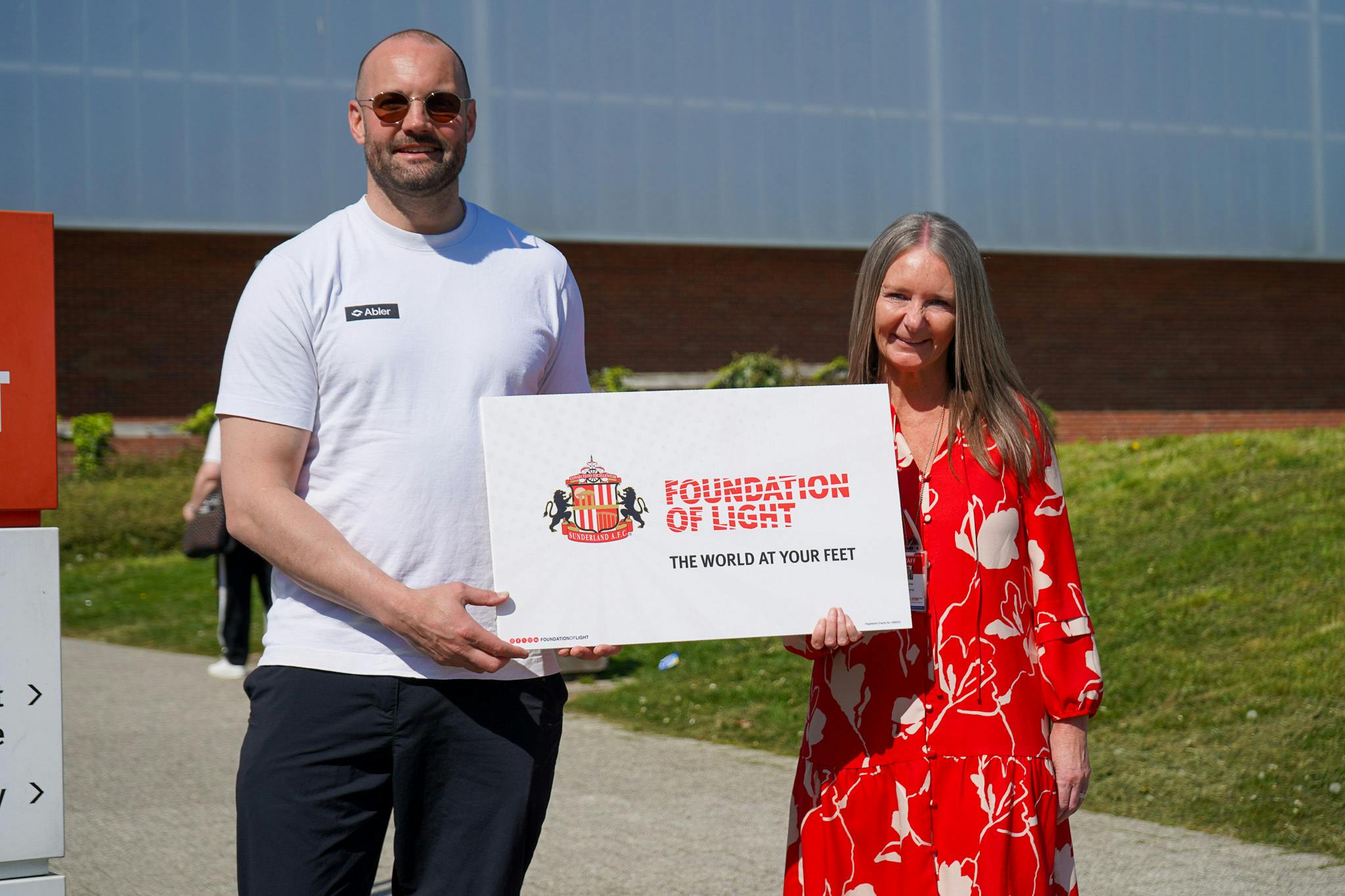 A man in a white t-shirt and a woman in a red floral dress hold a sign that reads "FOUNDATION OF LIGHT THE WORLD AT YOUR FEET". The sign also features the Sunderland AFC crest. They are standing outdoors in front of a building with a glass facade.