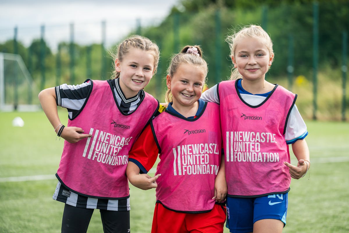 Three smiling young girls in Newcastle United Foundation training bibs on a field.