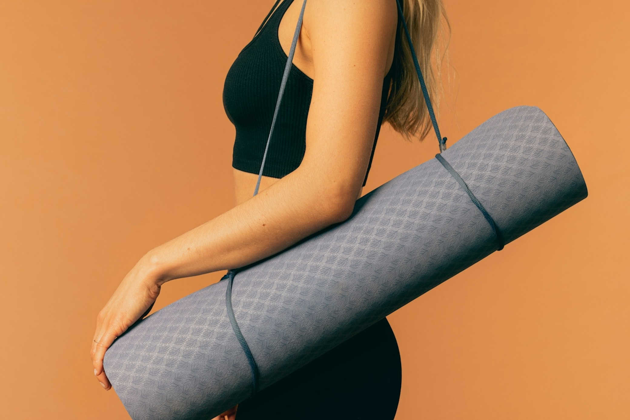 Woman in black athletic wear holding rolled gray yoga mat against orange background