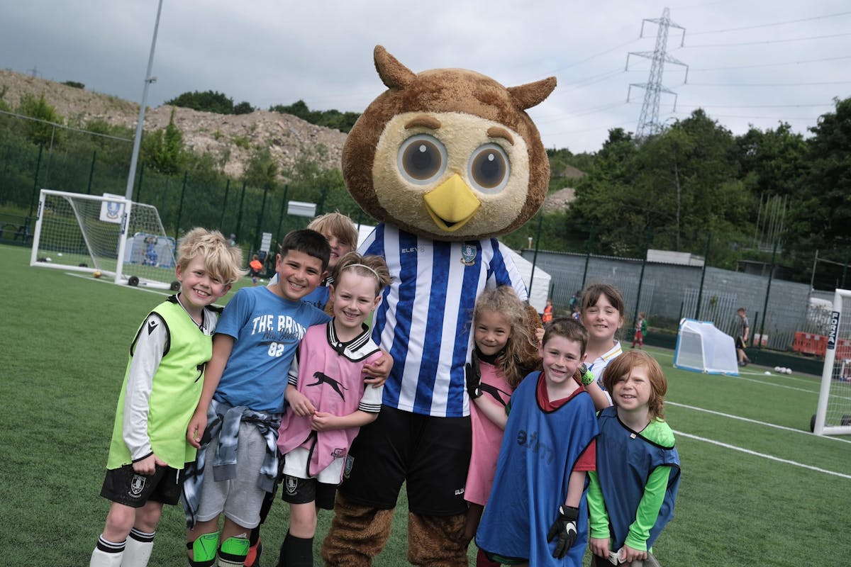 heffield Wednesday FC mascot Ozzie Owl poses with young football participants wearing colorful training bibs at the club's community training facility, showcasing youth engagement programmes