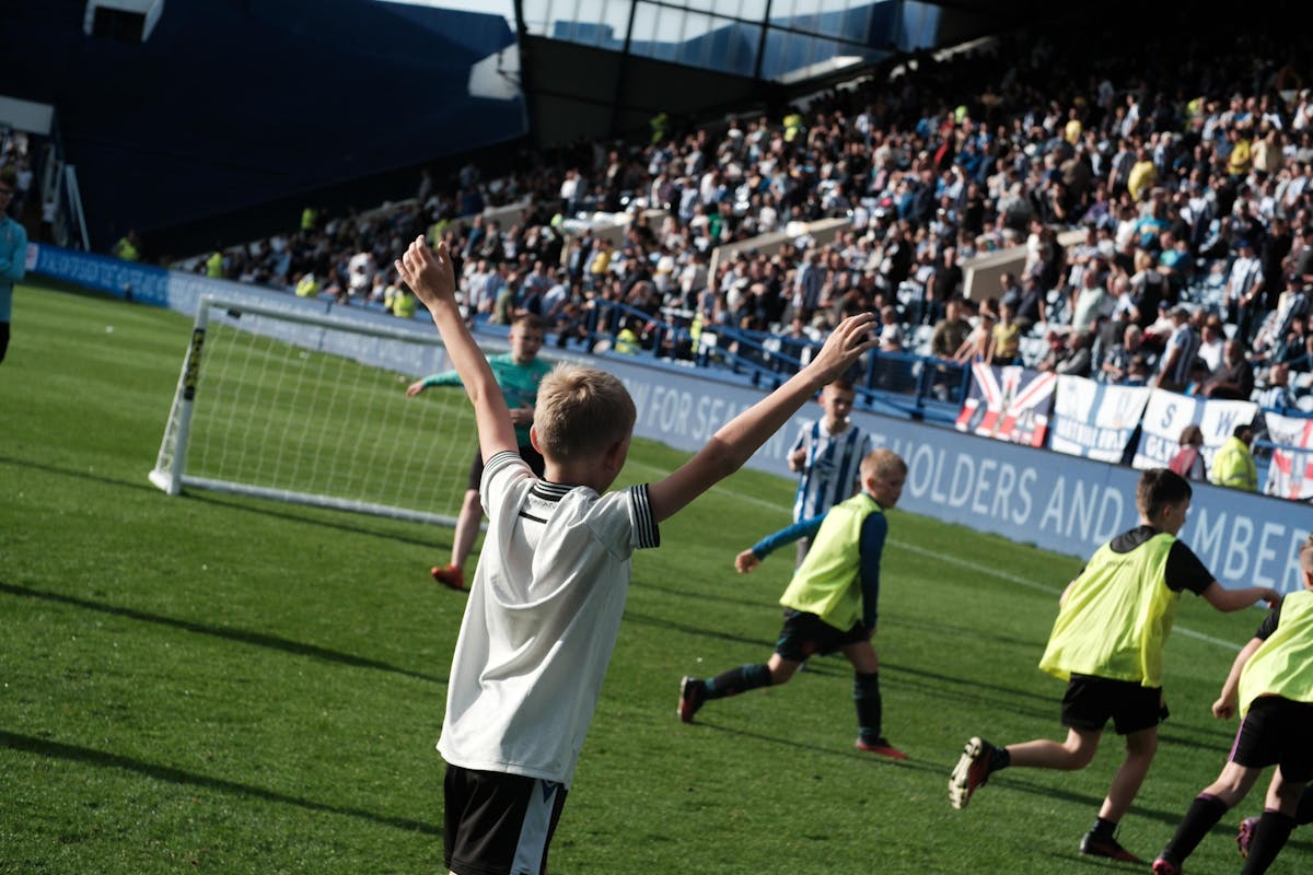 Young footballer celebrating with arms raised at Sheffield Wednesday's Hillsborough Stadium during a community programme match day experience, with packed stands visible in background