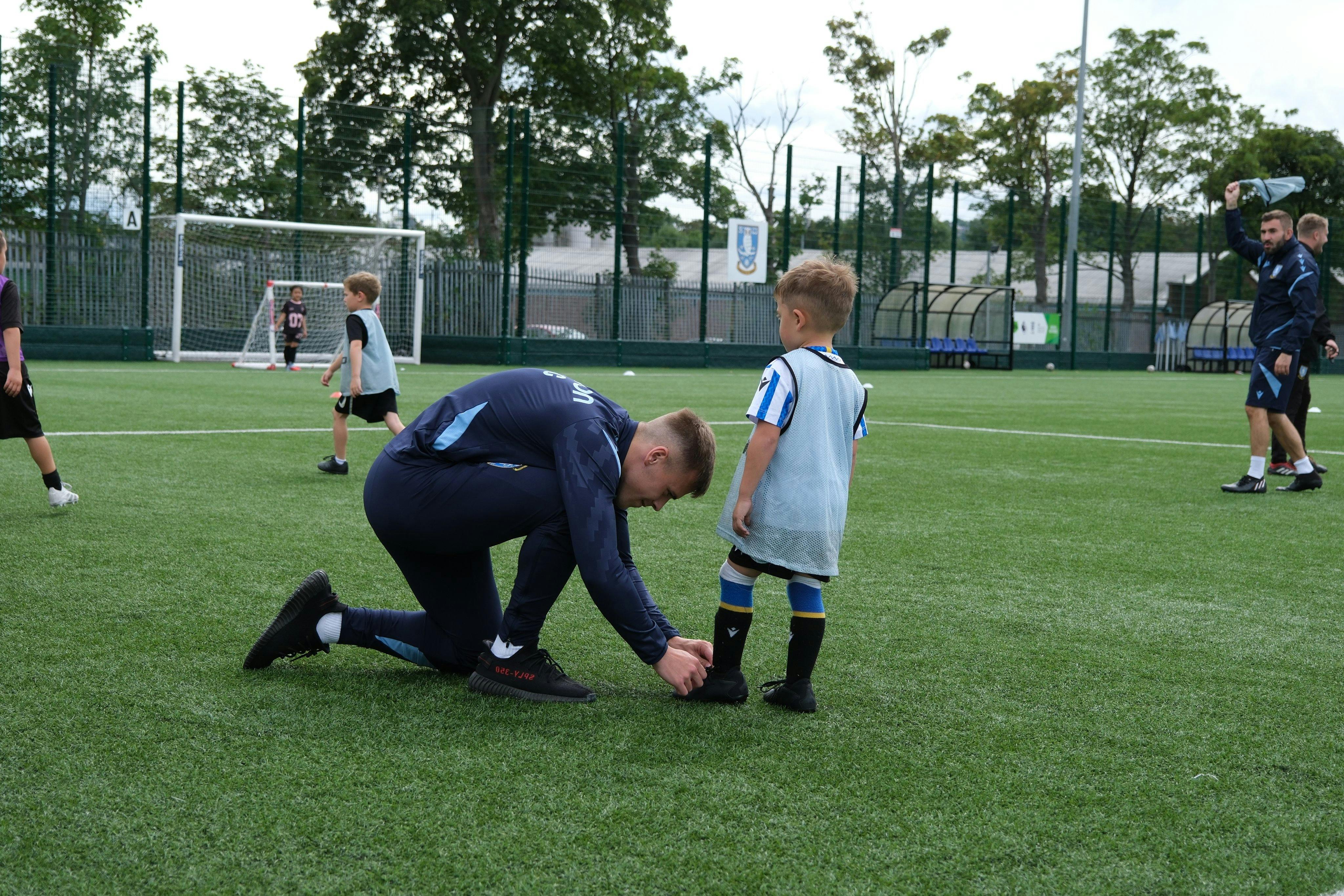 Sheffield Wednesday FC Community Programme coach helping young player tie football boots on artificial training pitch, demonstrating hands-on coaching and mentorship in youth development programmes